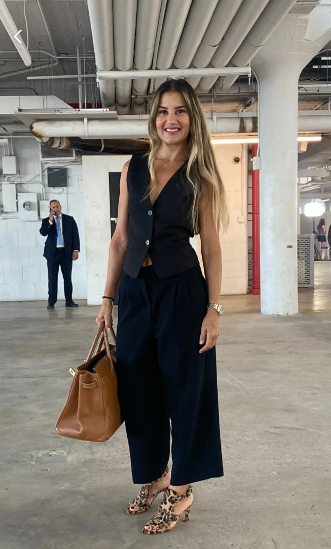 A woman with long blonde hair wearing a sleeveless black vest and wide-leg pants, holding a brown handbag, in an industrial parking garage with pipes overhead. She is smiling and wearing leopard-print high heels. A man in a suit talking on a phone is in the background.