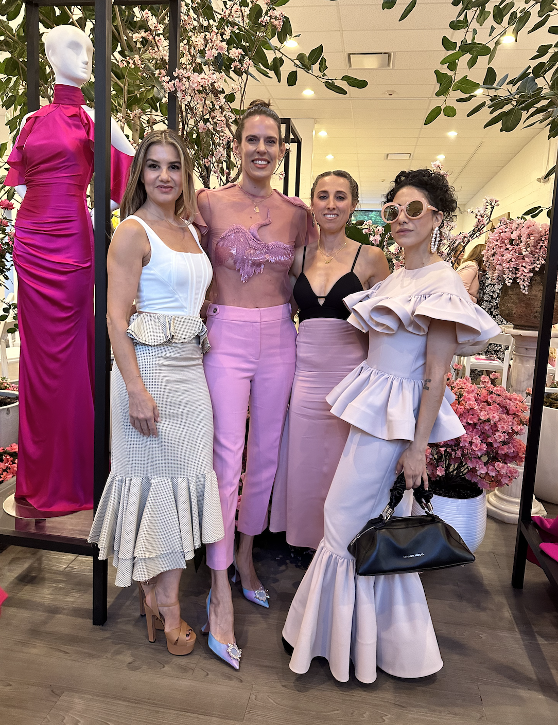 Four women standing together indoors, dressed in fashionable clothing, in front of pink flowers and greenery display.