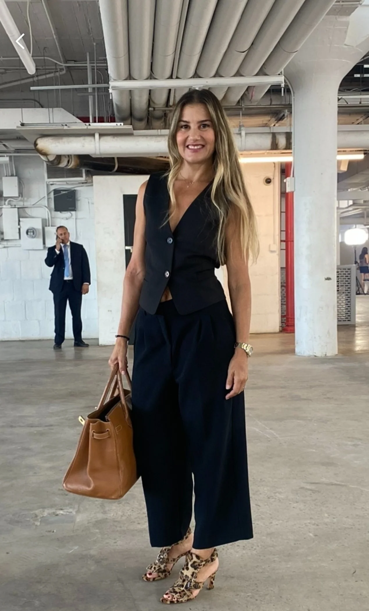 A woman in black pants and a sleeveless black vest stands in an industrial-looking space, holding a brown handbag, wearing leopard print high heels, smiling at the camera. A man in a suit is in the background talking on the phone.