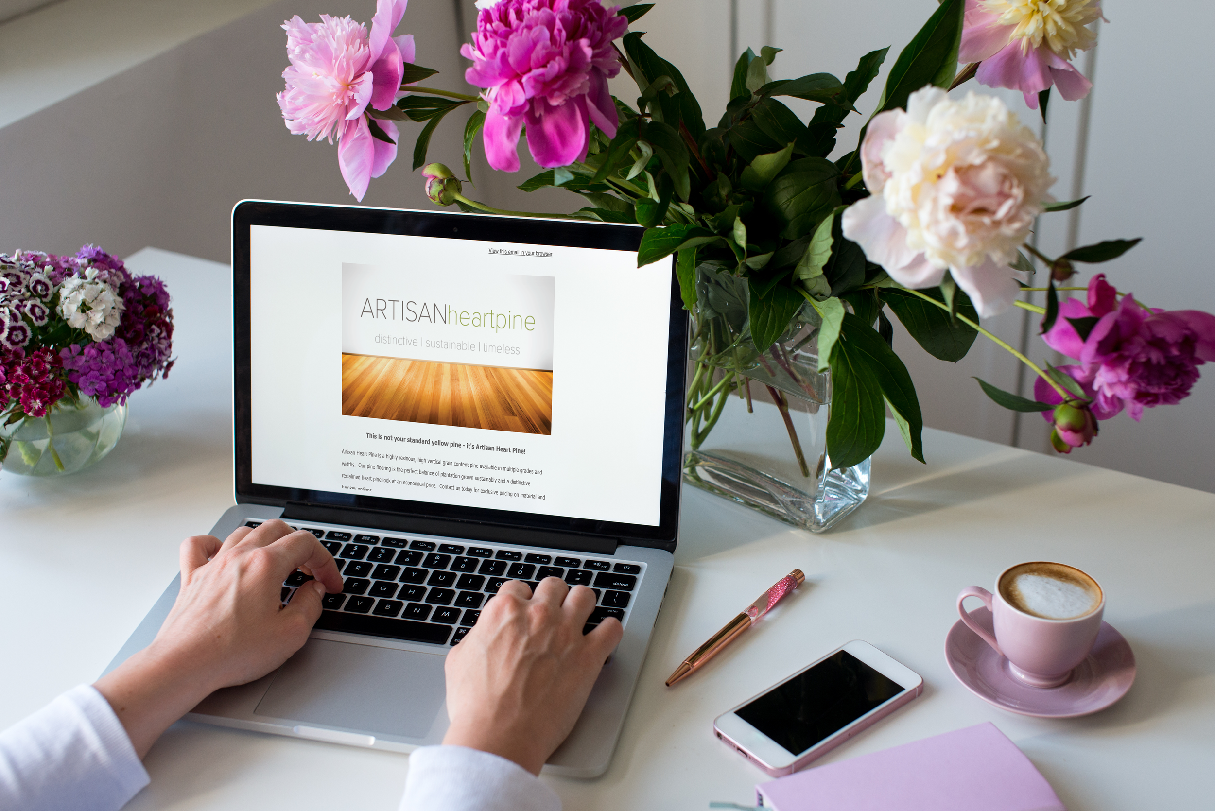 A person using a laptop on a white desk with a glass vase of pink, purple, and white flowers, a cup of coffee on a pink saucer, a smartphone, a pink notebook, and a pink and gold pen.