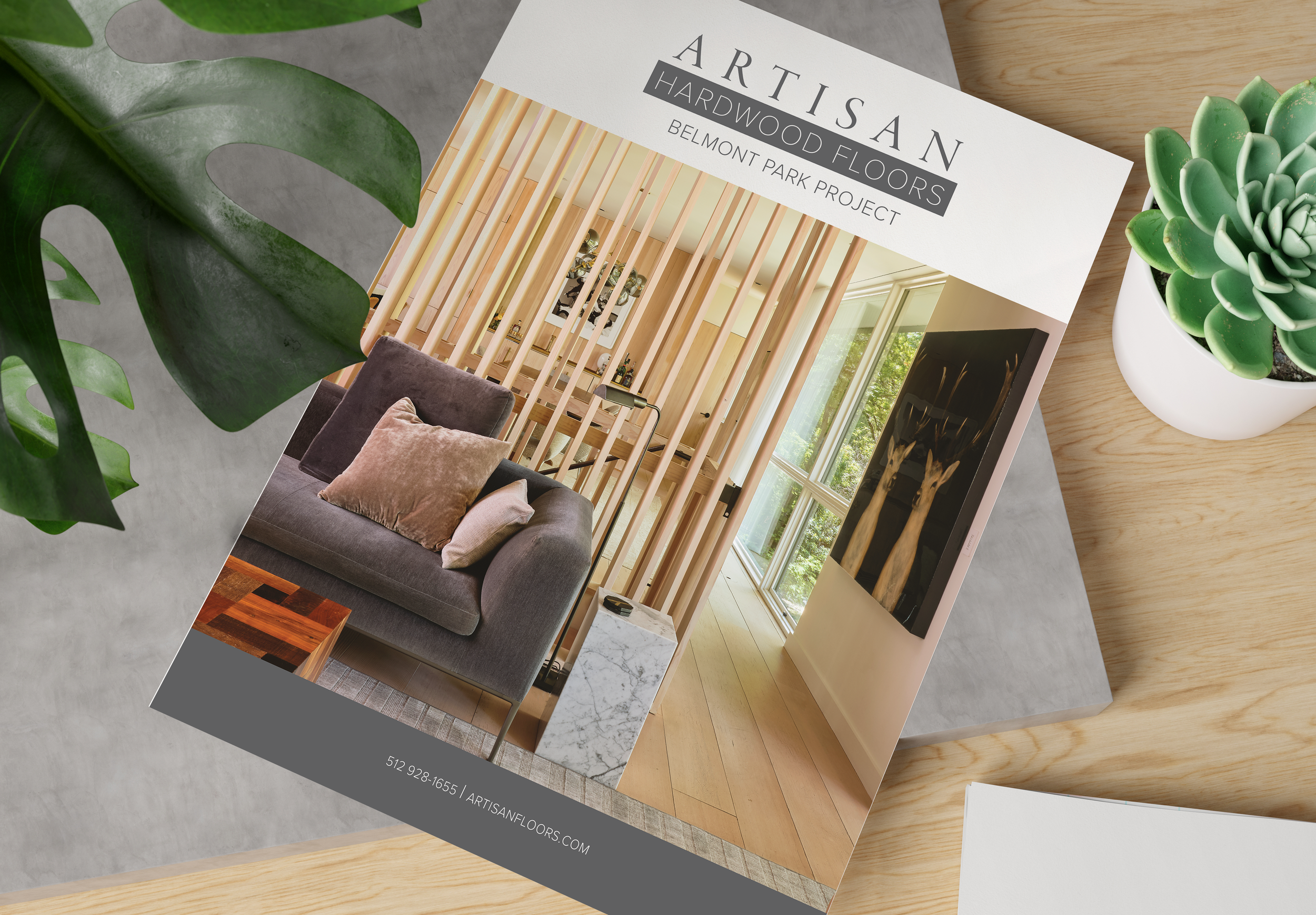 Brochure for Artisan Hardwood Floors featuring a modern living room with wood flooring, a dark grey sofa with pillows, a marble side table, and a large window with natural light, placed on a light wooden table with a potted succulent plant.