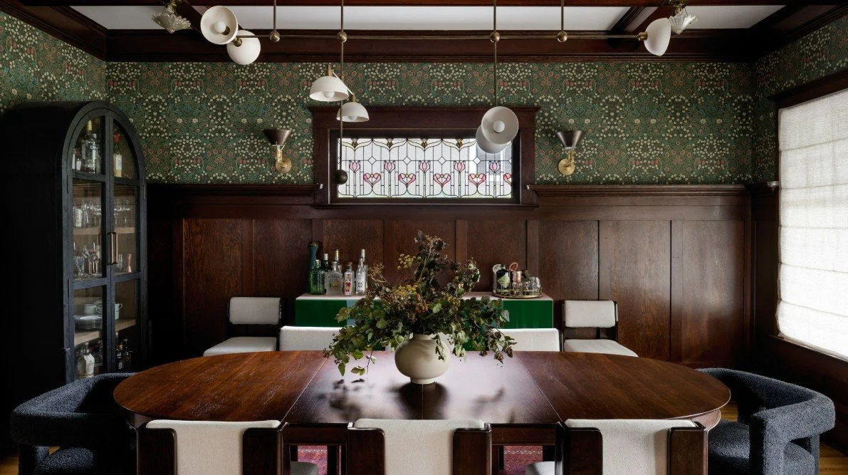 A dining room with a large wooden table, a floral centerpiece vase, a window with stained glass, and a dark cabinet filled with glassware in the corner.