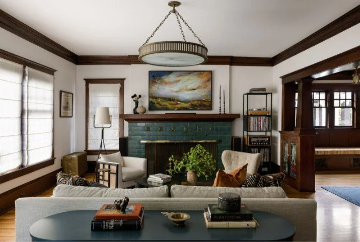 Living room with white walls, dark wood trim, a fireplace with a colorful landscape painting above, and a mix of furniture including a white sofa, armchairs, and a blue side table. There is a circular chandelier hanging from the ceiling, and a plant on the coffee table.