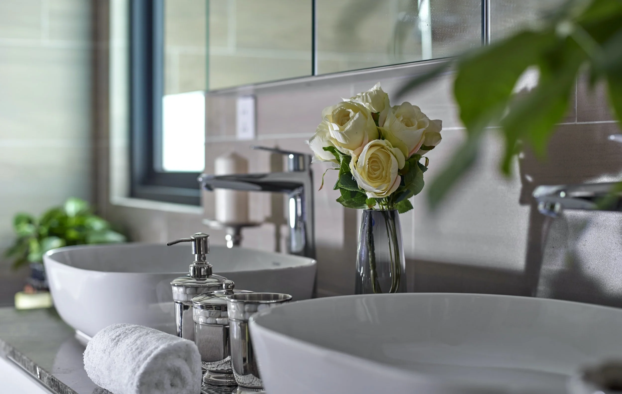 A bathroom with a white sink, a silver faucet, and a rolled white towel on the counter. A glass vase with white roses is placed at the sink. There is a window in the background with a view outside, and green leaves slightly obscure the right side of the image.