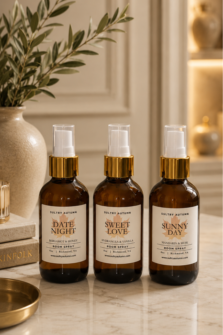 Three amber glass spray bottles with white spray nozzles and gold accents, labeled 'Date Night', 'Sweet Love', and 'Sunny Day', sit on a marble countertop next to a beige vase with green foliage and a stack of books.