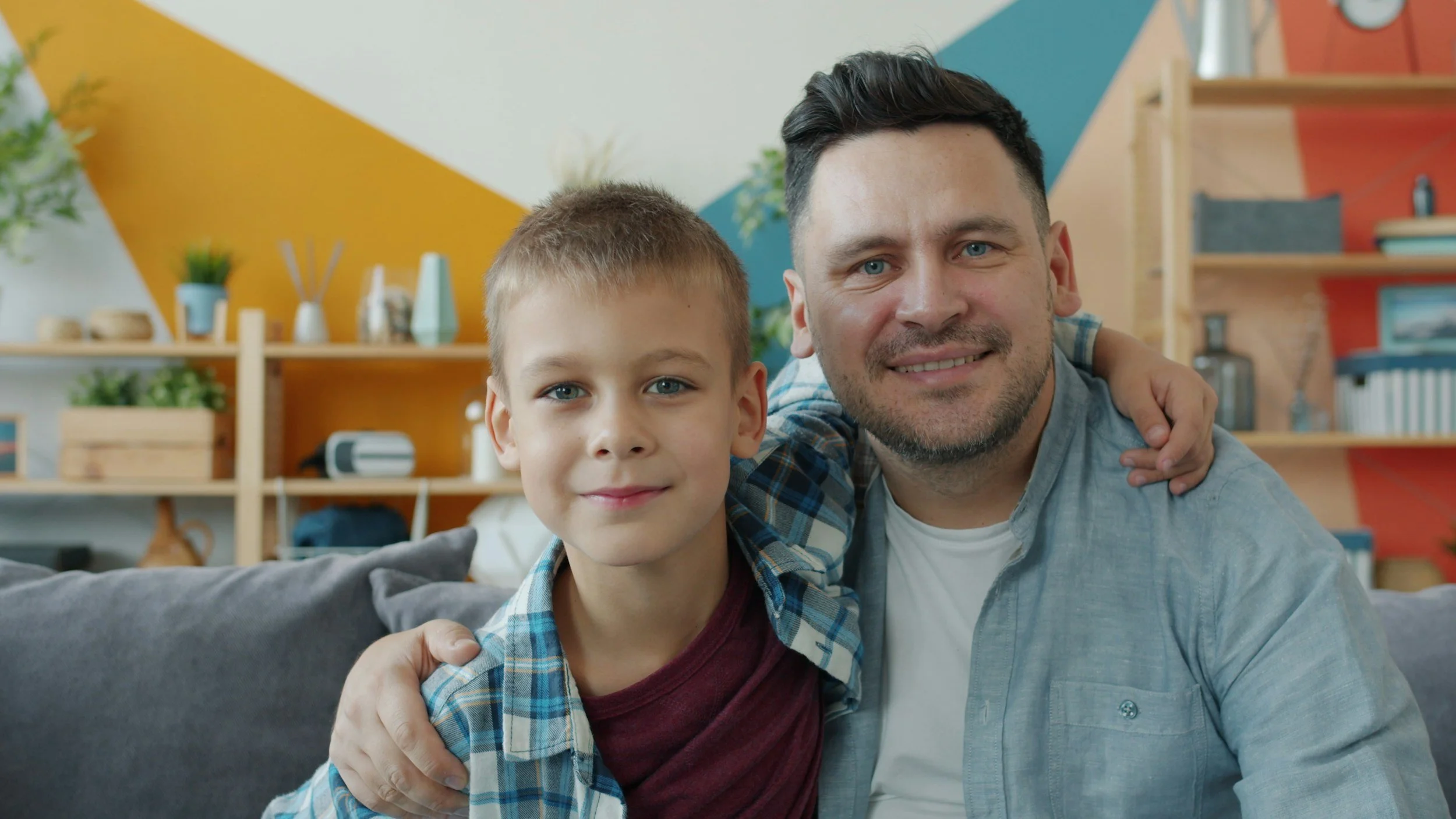 A man with dark hair and a beard, and a young boy with short blond hair and blue eyes, smiling and sitting close together on a gray couch in a colorful modern living room.