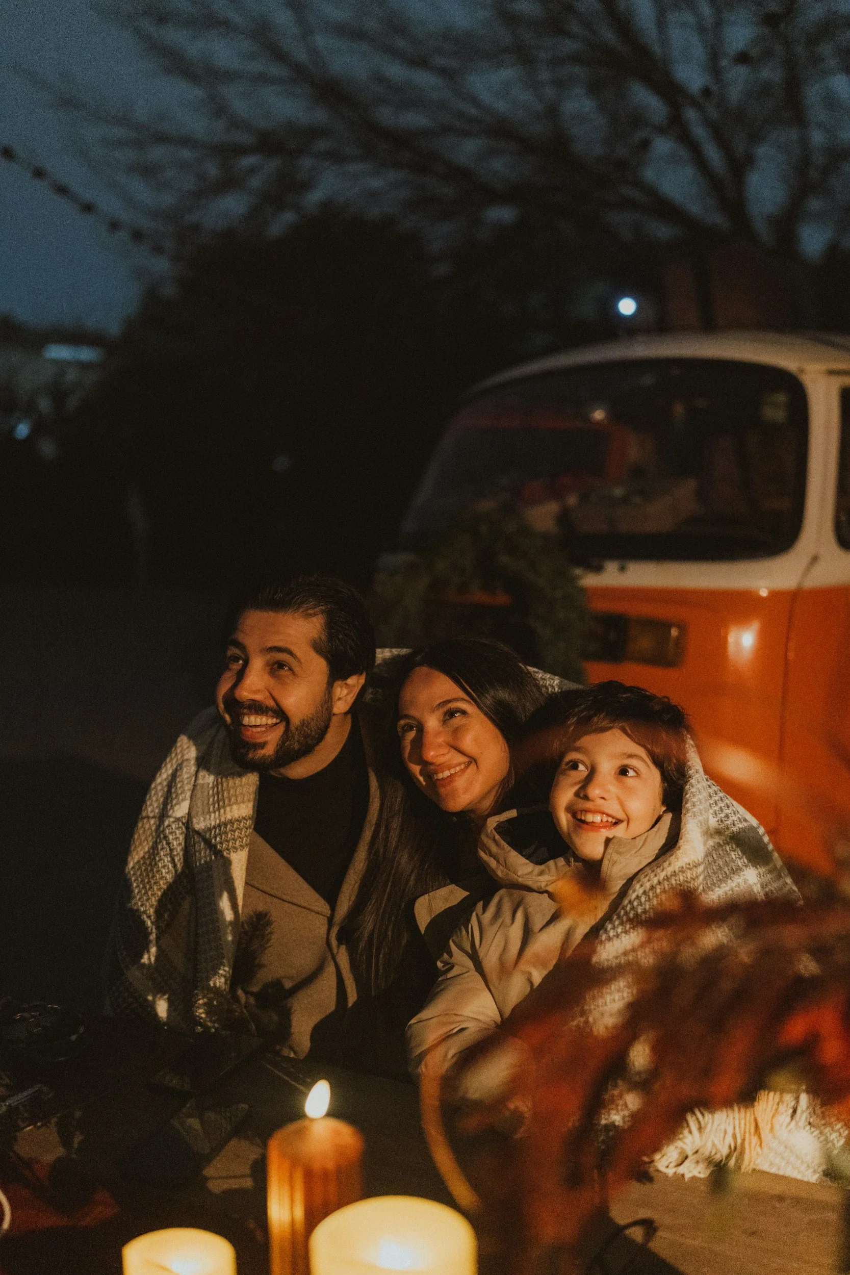 Family enjoying a cozy evening gathering by candles outside at night, smiling and looking happy.