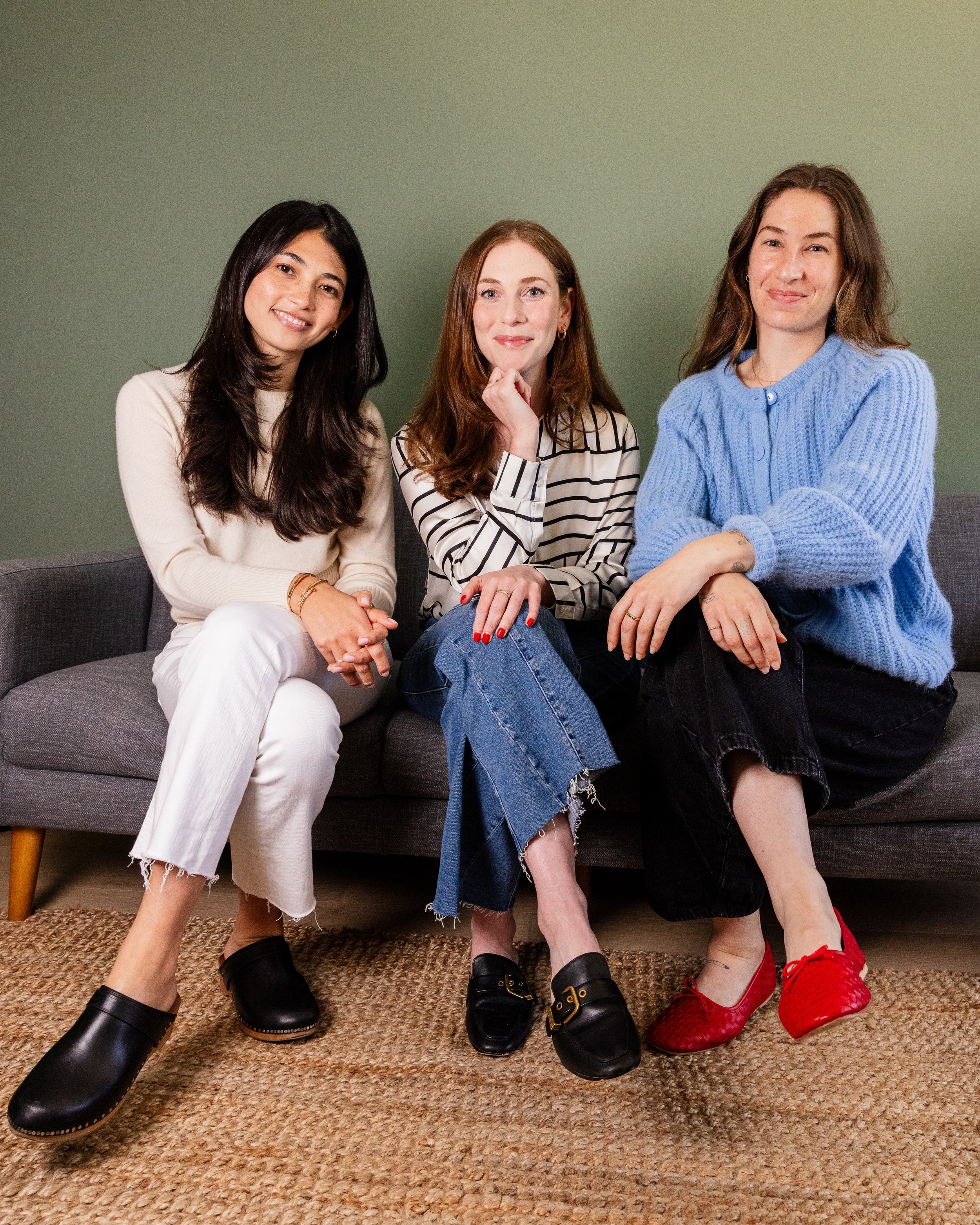 Three women sitting on a gray sofa against a green wall, smiling at the camera. The woman on the left is wearing a cream-colored sweater and white pants, the woman in the middle is wearing a striped blouse and jeans, and the woman on the right is wearing a blue sweater and black pants.
