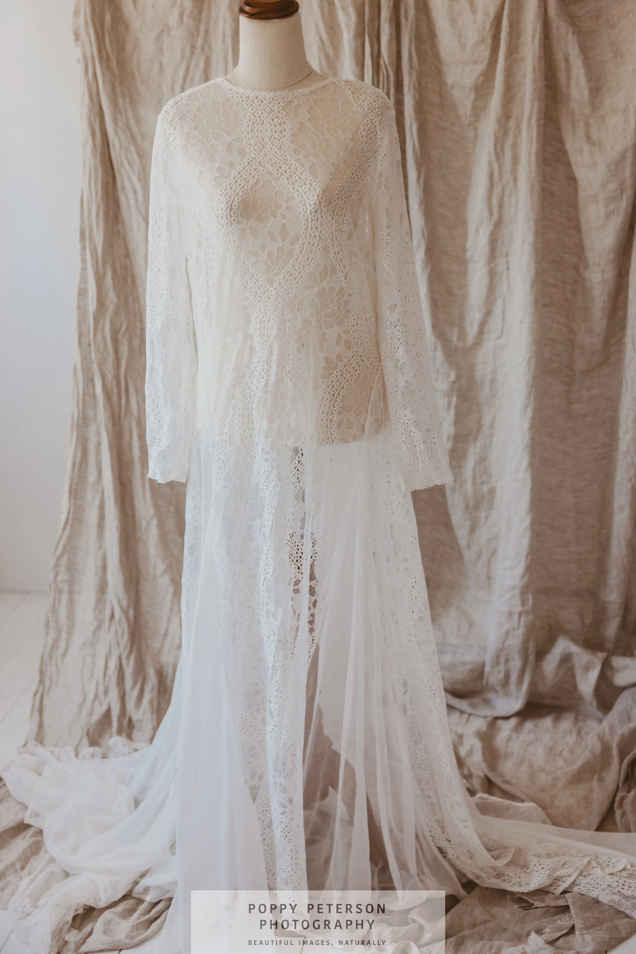 A white lace wedding dress on a mannequin with intricate lace details, long sleeves, and a flowing train, set against beige fabric backdrop.