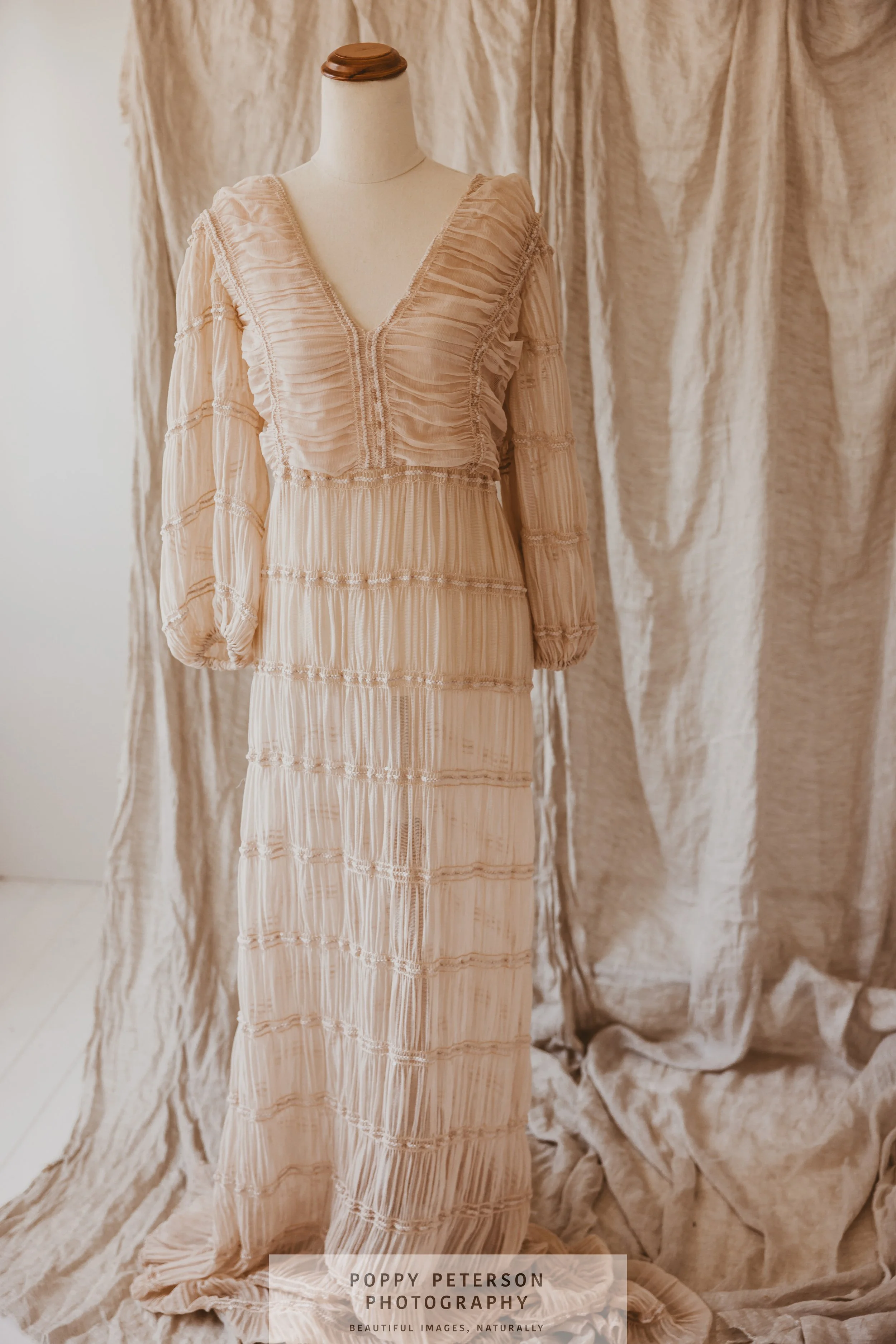 Beige vintage-style dress on display, featuring intricate pleats and lace detailing, against a backdrop of textured beige fabric.