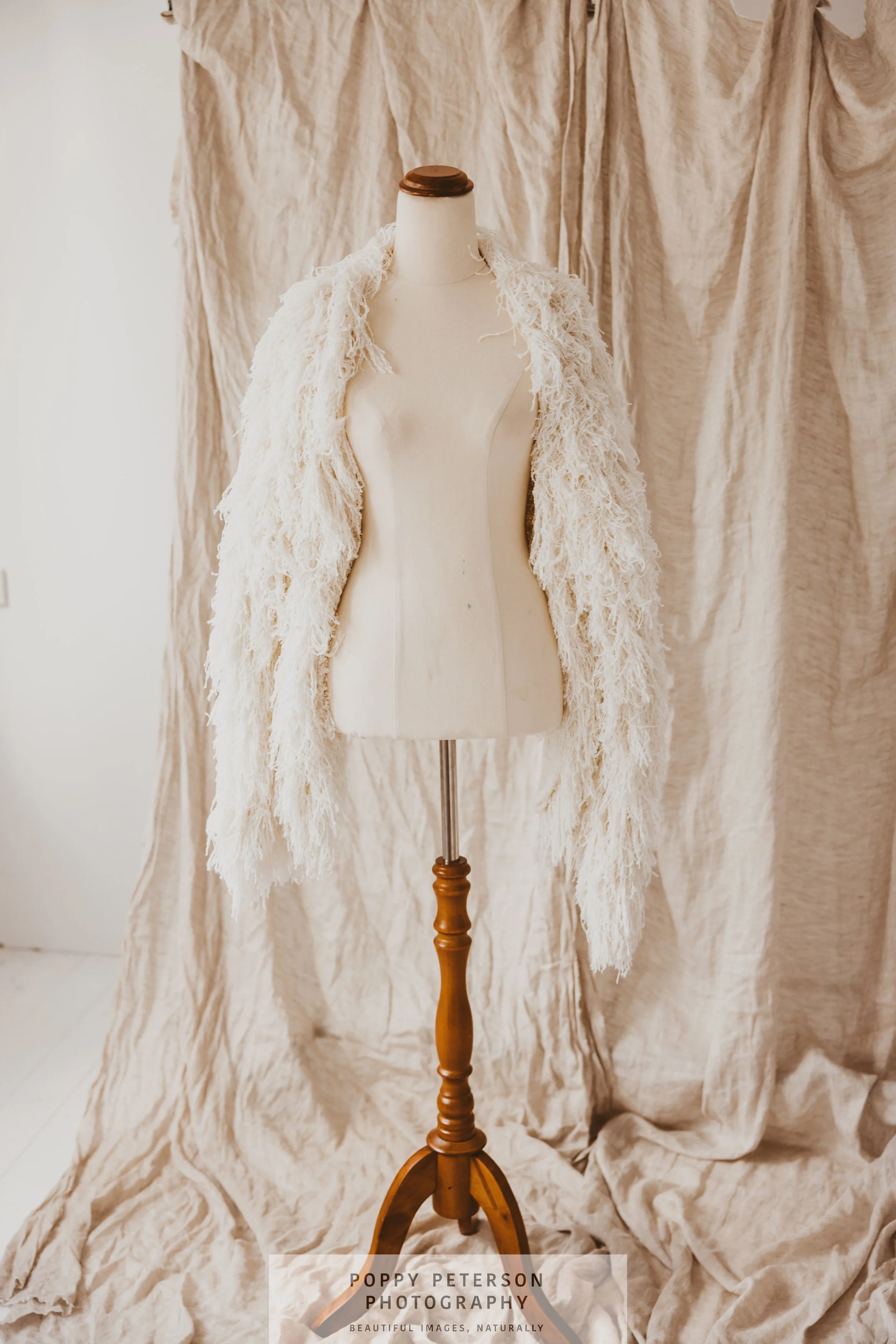 A mannequin wearing a fluffy, cream-colored textured cardigan over a dress form, with a wrinkled beige fabric backdrop.