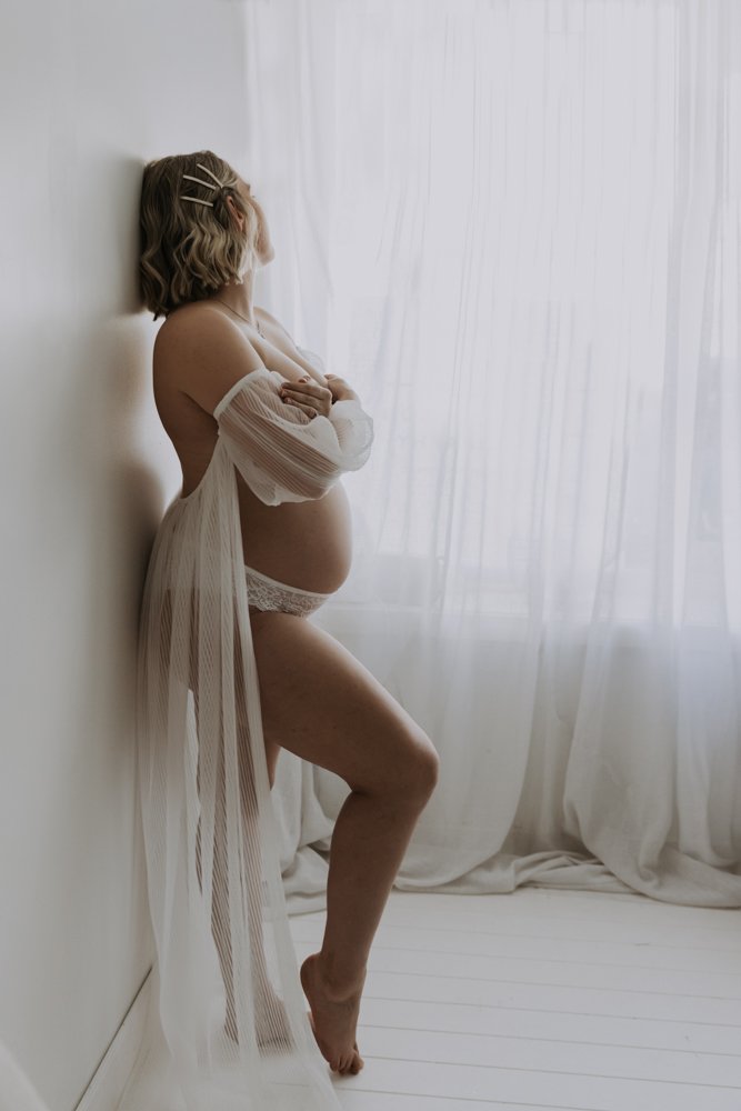 Pregnant woman in white sheer robe standing by a window, arms crossed, with soft natural light filtering through curtains.