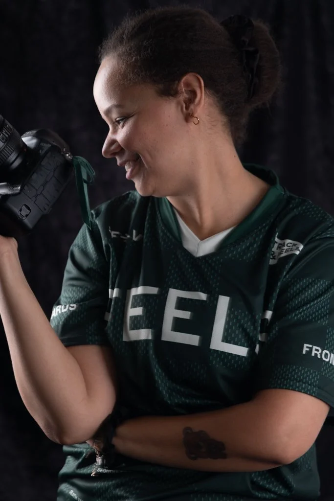 Young woman with curly hair in a bun, wearing a dark green sports jersey, looking through a camera, smiling, with a dark background.