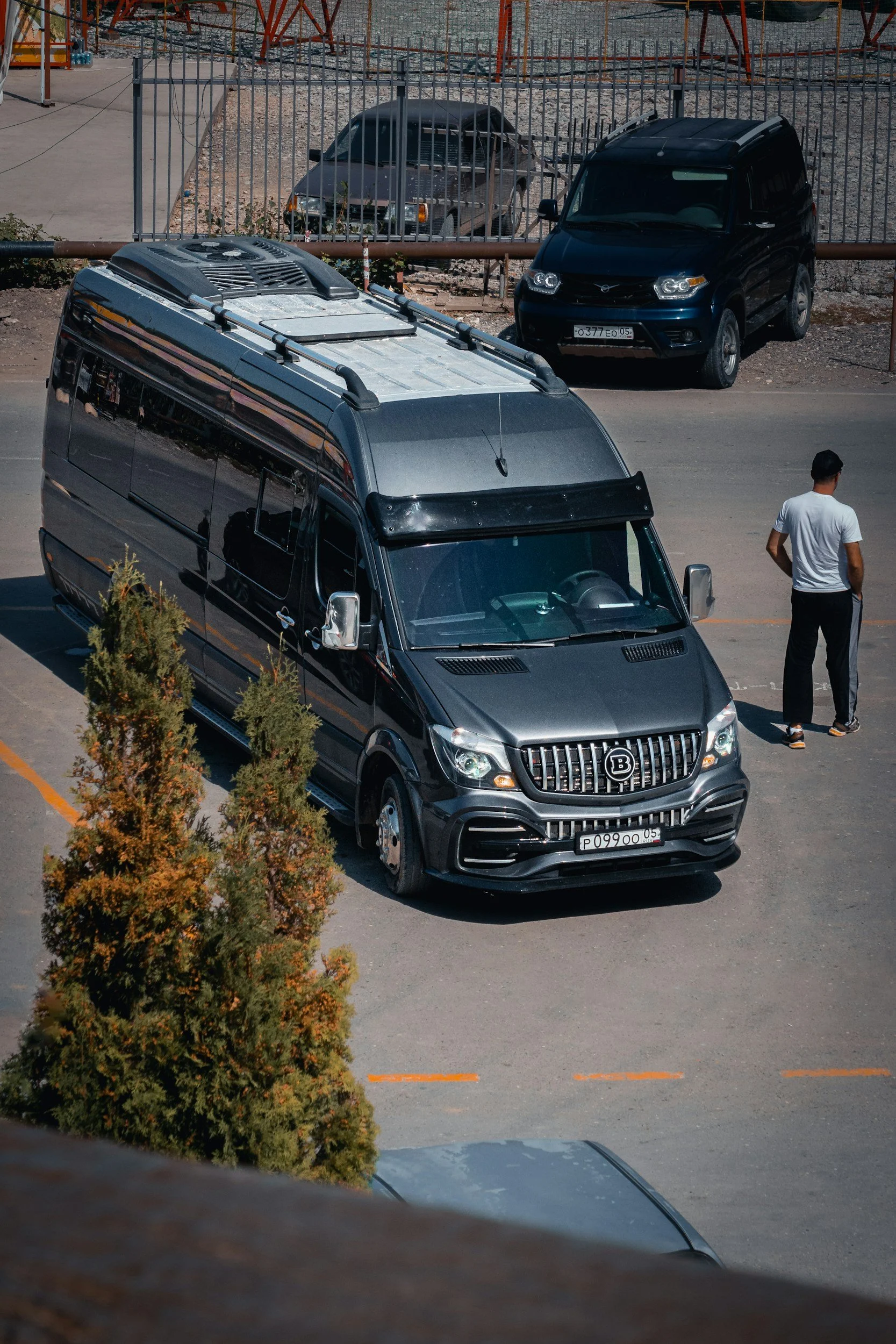 A parking lot with various parked cars, including a black van in the foreground and a dark green car and a gray car behind a fence. A person wearing a white t-shirt and black pants stands near the black van, and there are small bushes along the parking area.