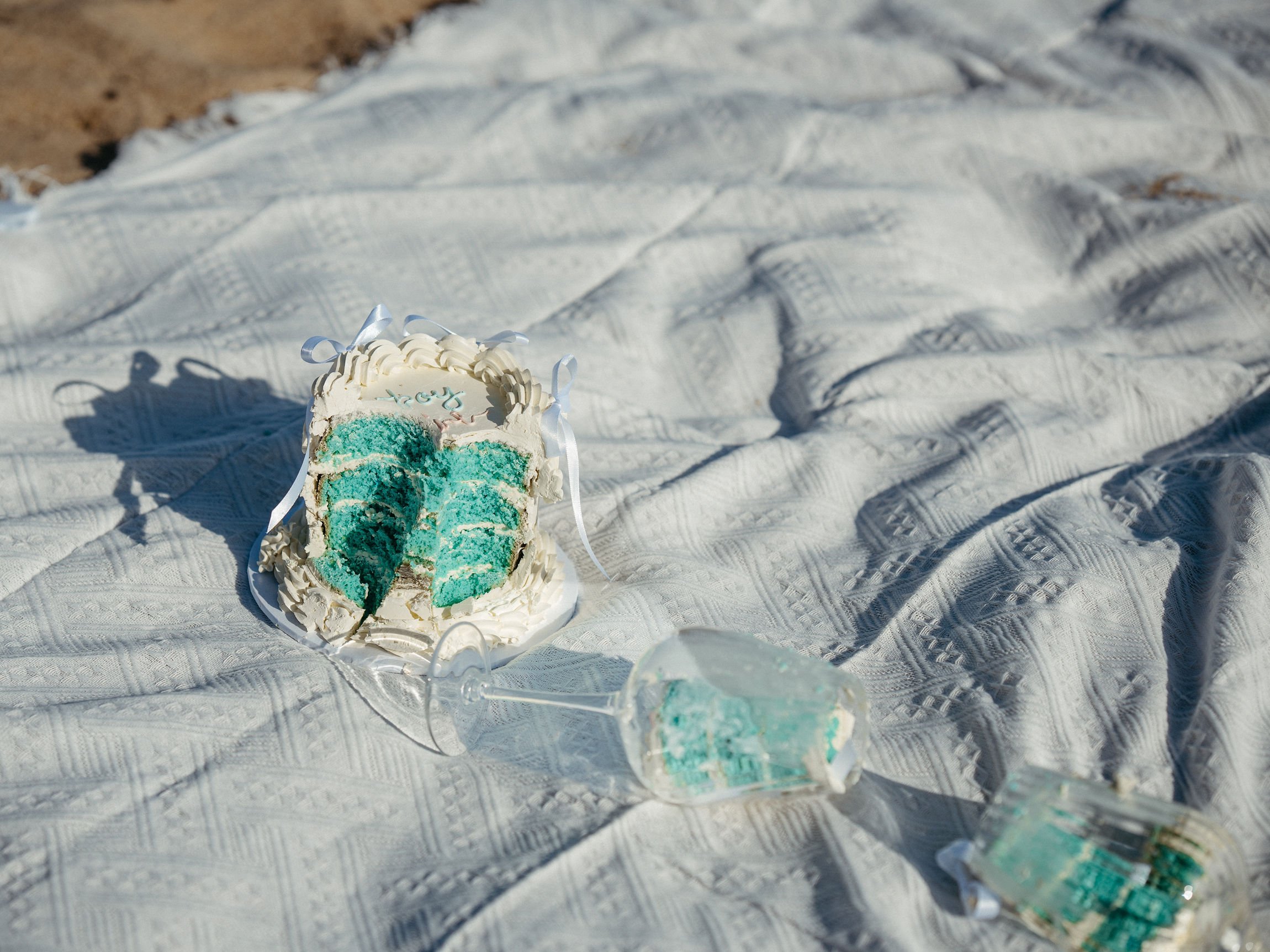 A partially eaten turquoise cake with white frosting and ribbon decorations on a white textured cloth.