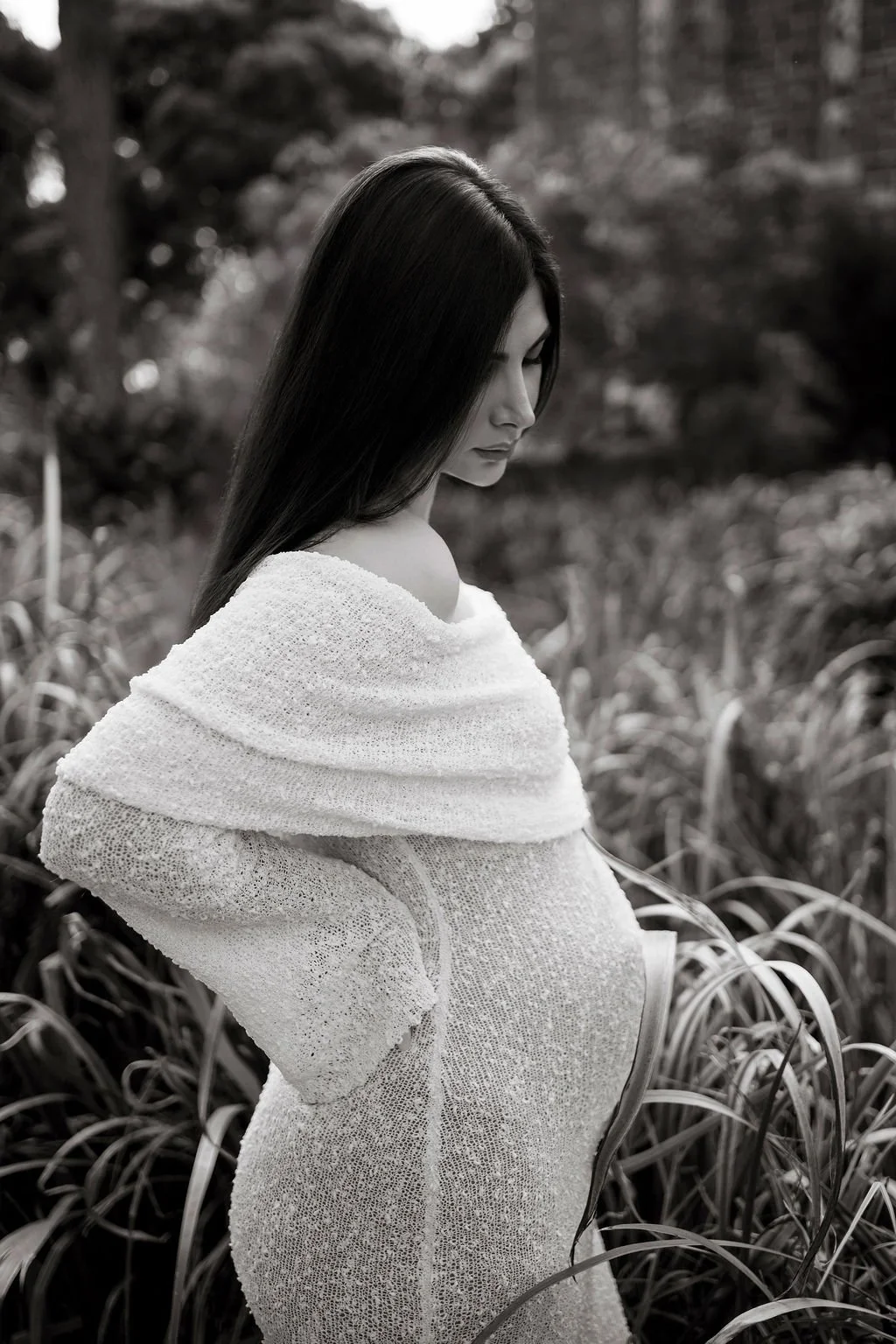 A woman with long dark hair standing outdoors, wrapped in a light-colored, textured shawl and dress, looking down with a serene expression, surrounded by tall grasses and trees in a natural setting.