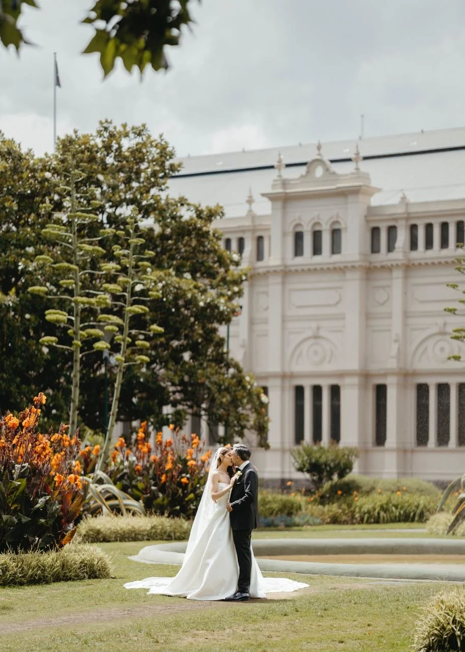 A bride and groom in wedding attire embracing and kissing outdoors on a grassy area with flowers, trees, and a historic building in the background.