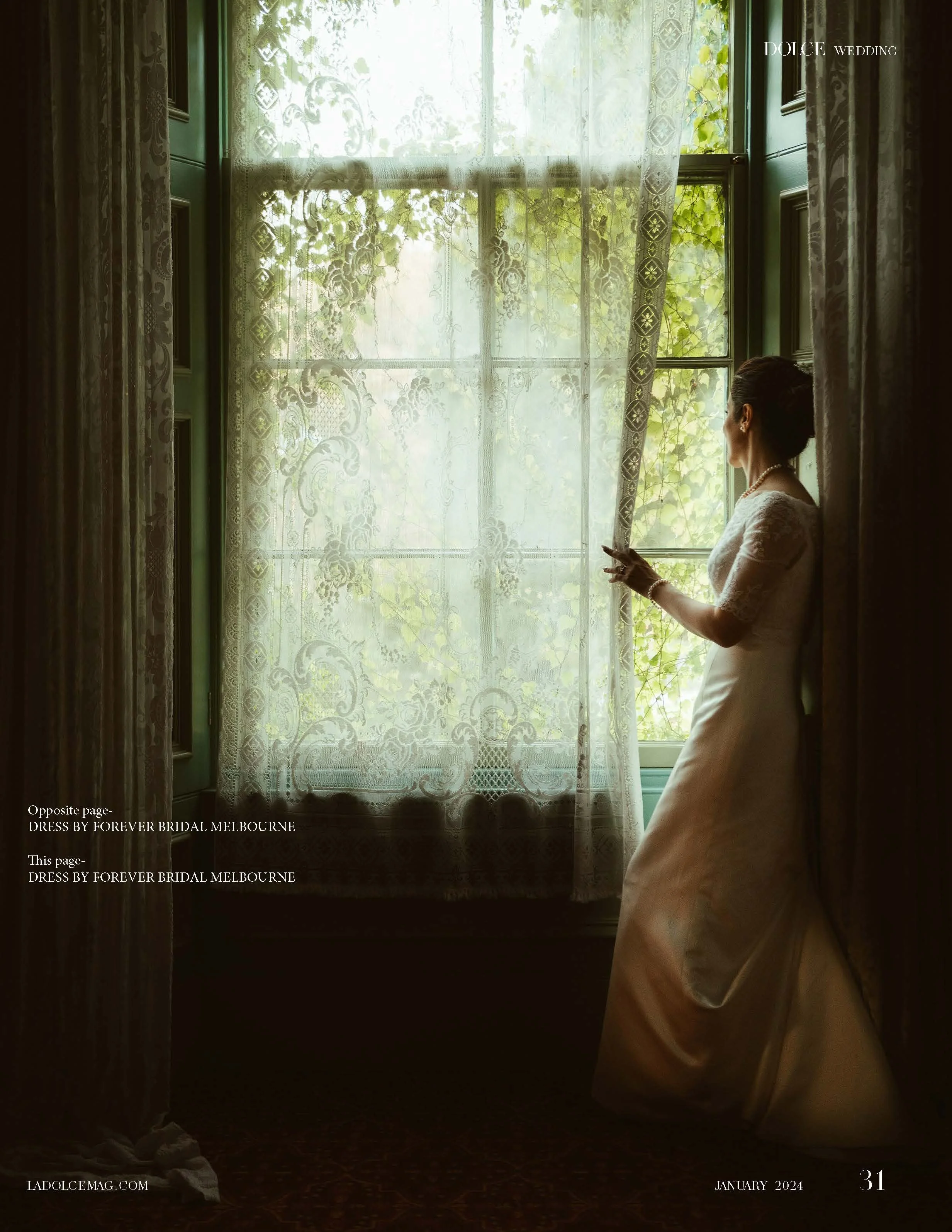 A woman in a white wedding dress standing by a window with lace curtains, looking outside at a vine-covered outdoor scene.