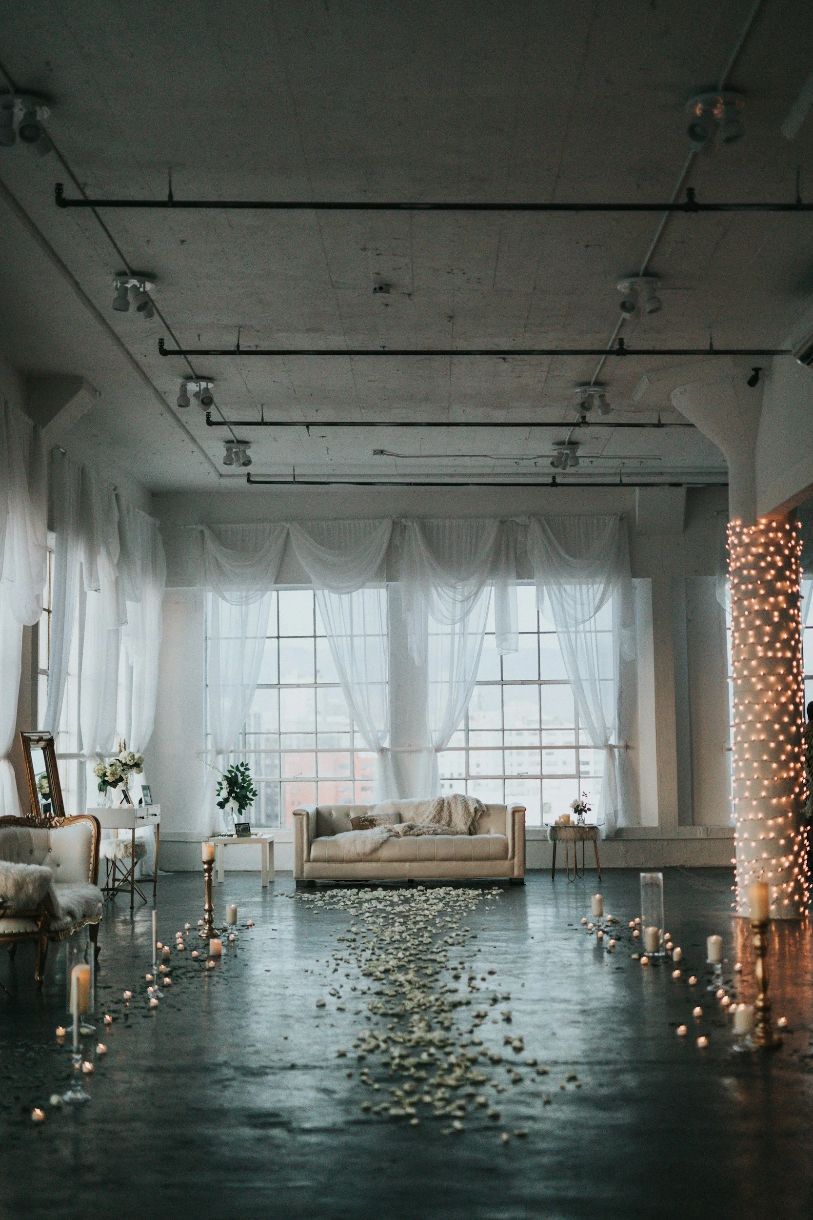Empty indoor venue decorated for a wedding with white curtains, a cream sofa, candles, and flower arrangements, illuminated by soft natural light from large windows.