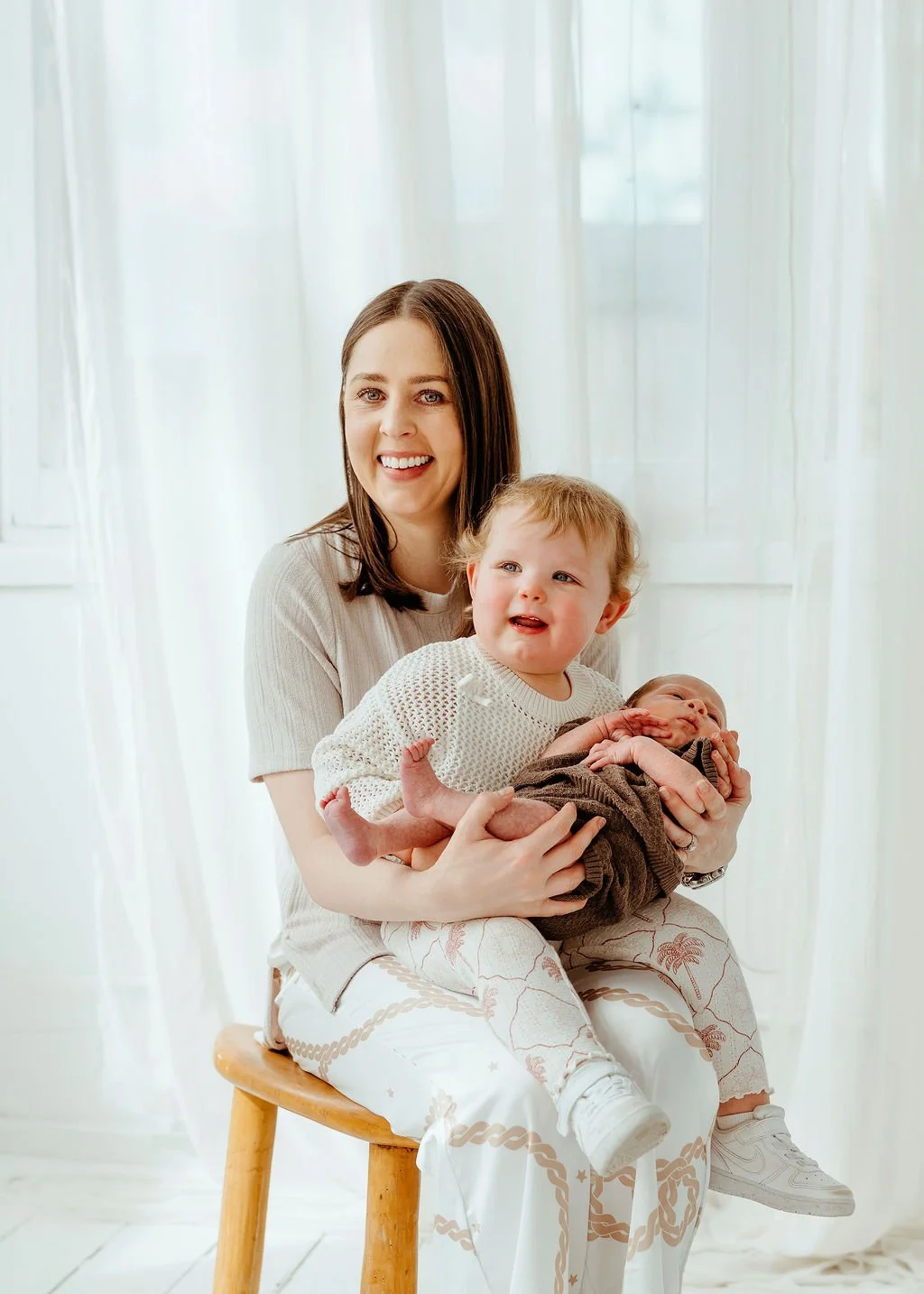 A smiling woman sitting on a wooden stool holding a young boy and a baby, with soft white curtains in the background.