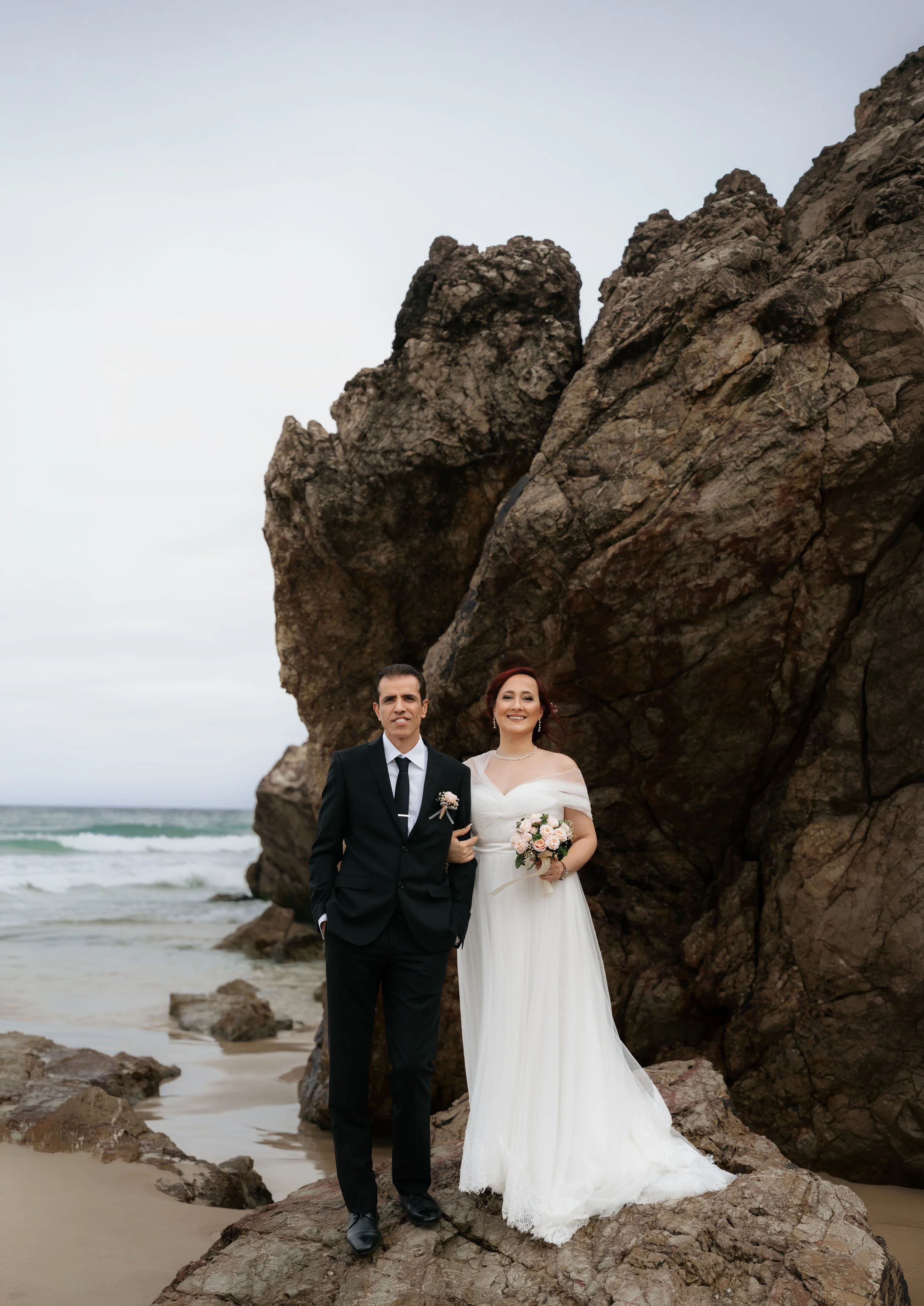 A newlywed couple holding hands and smiling outdoors. The groom is in a black tuxedo with a bow tie, and the bride is in a white wedding dress holding a bouquet of flowers. There are trees and a gazebo in the background.