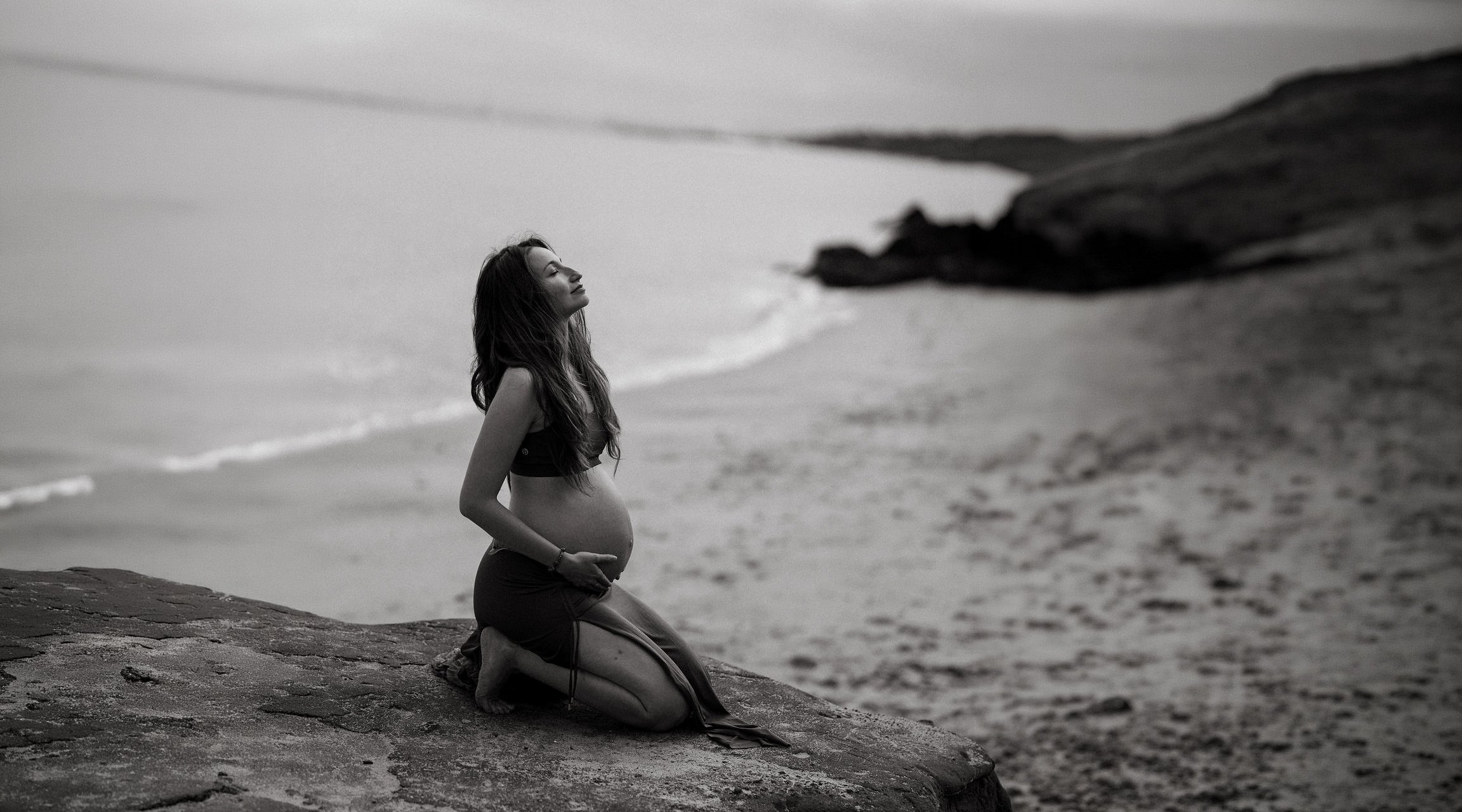 A pregnant woman kneels on a rock at the beach with her eyes closed and a peaceful smile, facing the ocean in black and white.
