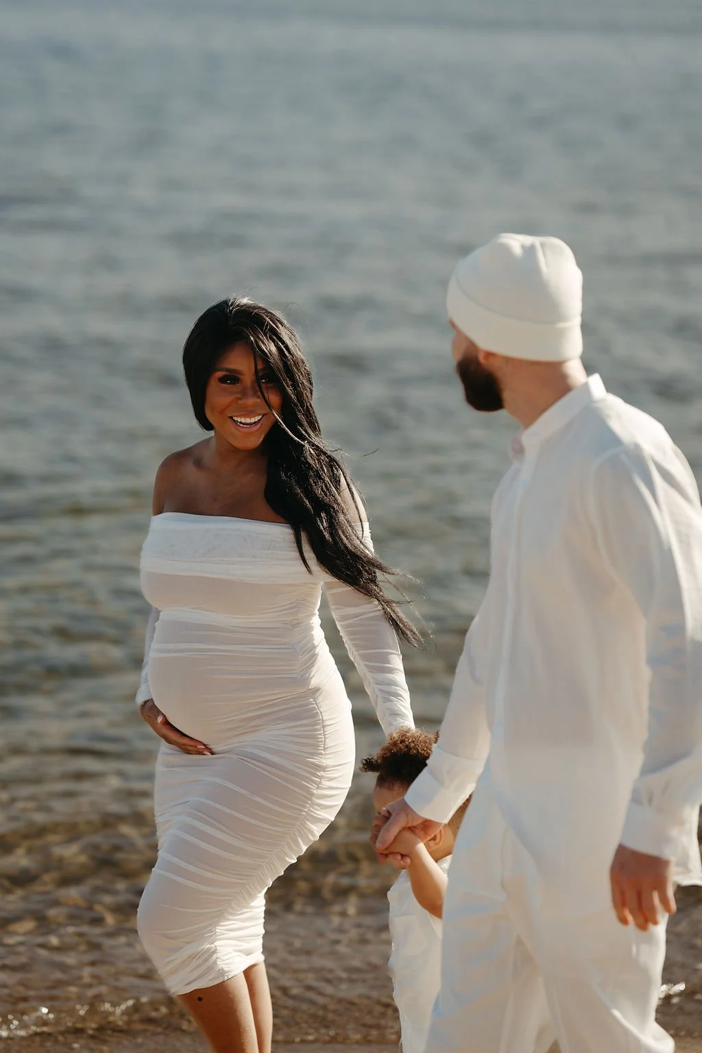 A pregnant woman in a white off-the-shoulder dress and a man in white clothing with a beanie, holding the hand of a young child at the beach.