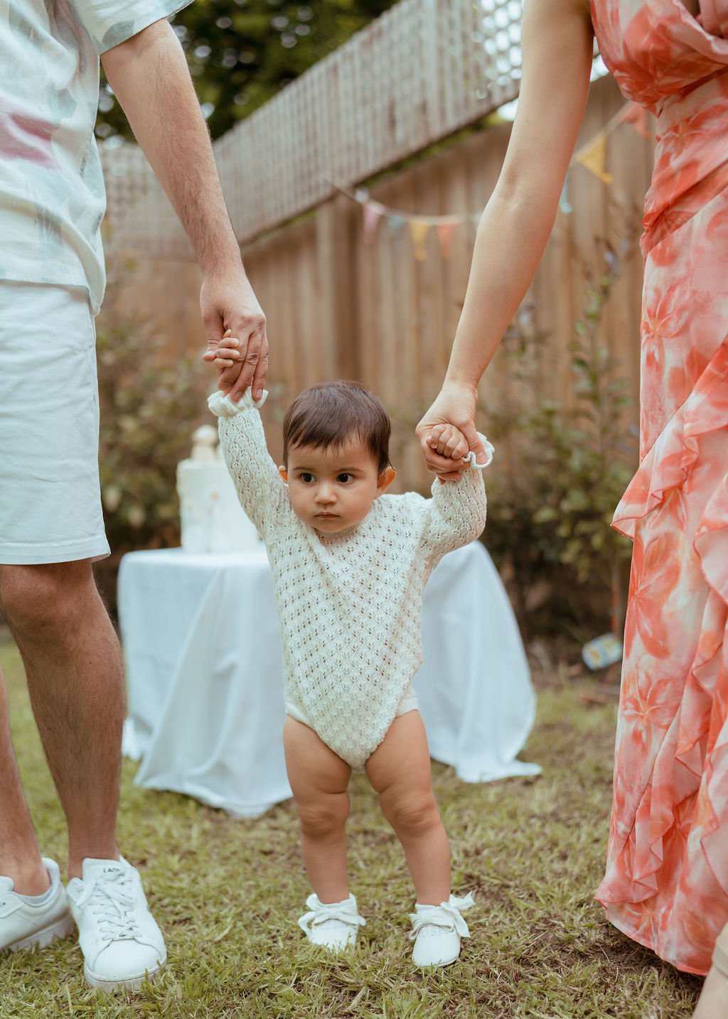 A young child in a cream-colored sweater and diaper shoes holds hands with two adults at an outdoor celebration, with a cake in the background.