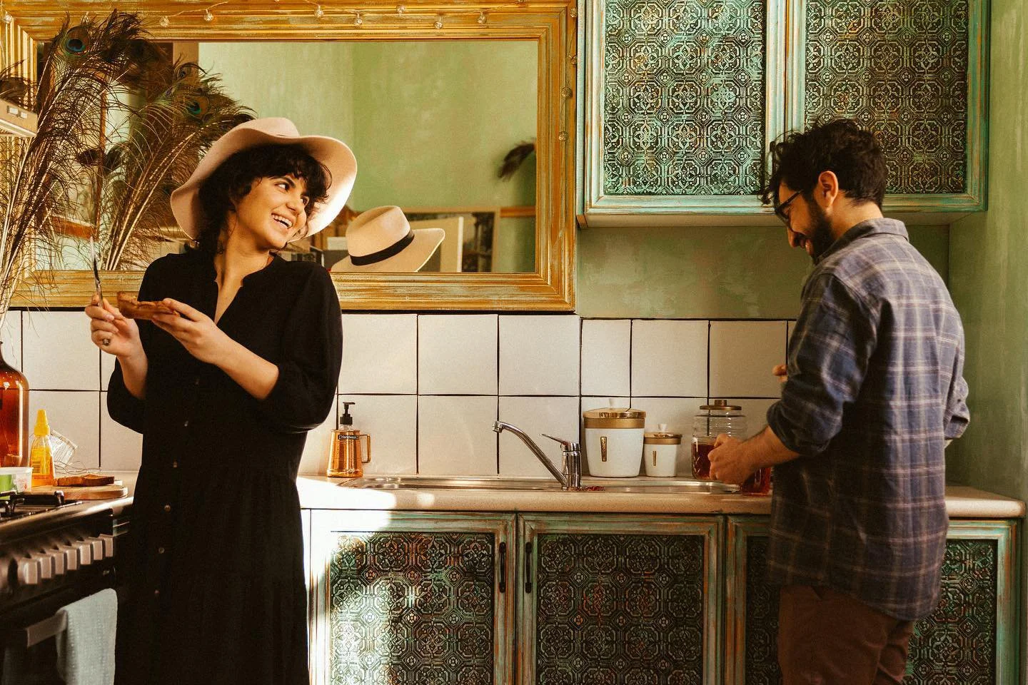 A woman in a black dress and large white hat holding a slice of pizza, smiling and looking at a man in a plaid shirt. The man is standing at the kitchen counter, preparing food or drink. The kitchen has vintage-style green and patterned cabinets, a large mirror, and various kitchen items on the countertop.