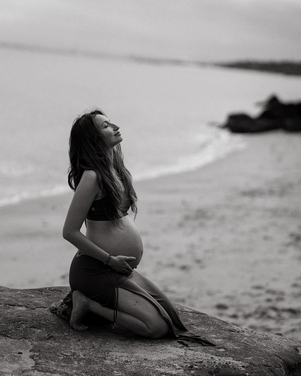A pregnant woman with long hair kneeling on a beach rock with her eyes closed, holding her belly, during sunset or sunrise.