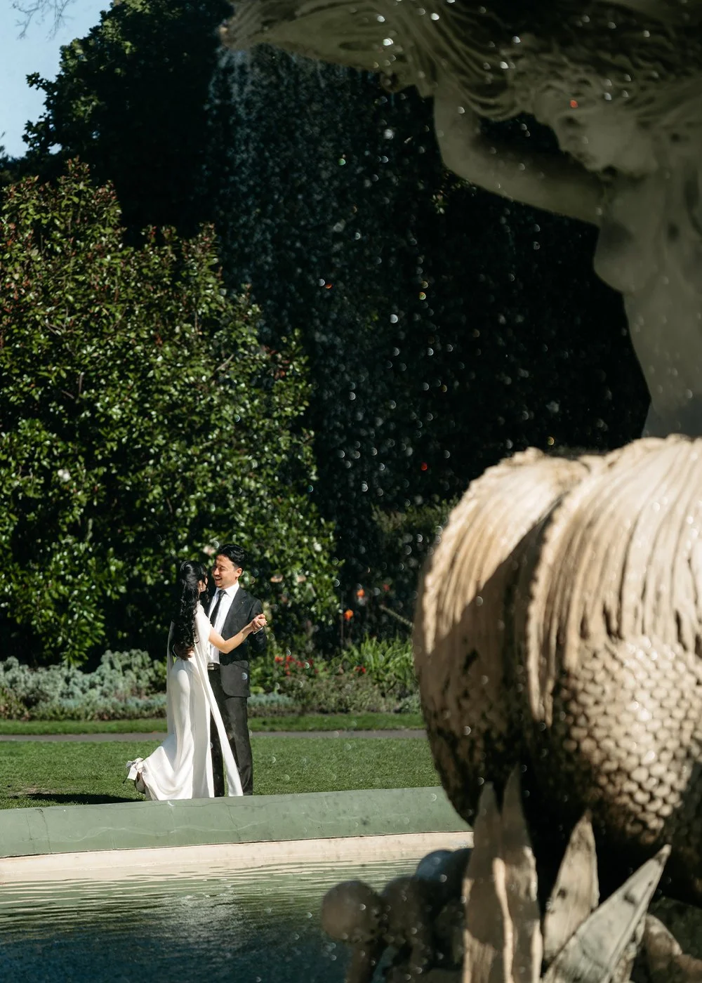 A newlywed couple holding hands and smiling outdoors. The groom is in a black tuxedo with a bow tie, and the bride is in a white wedding dress holding a bouquet of flowers. There are trees and a gazebo in the background.