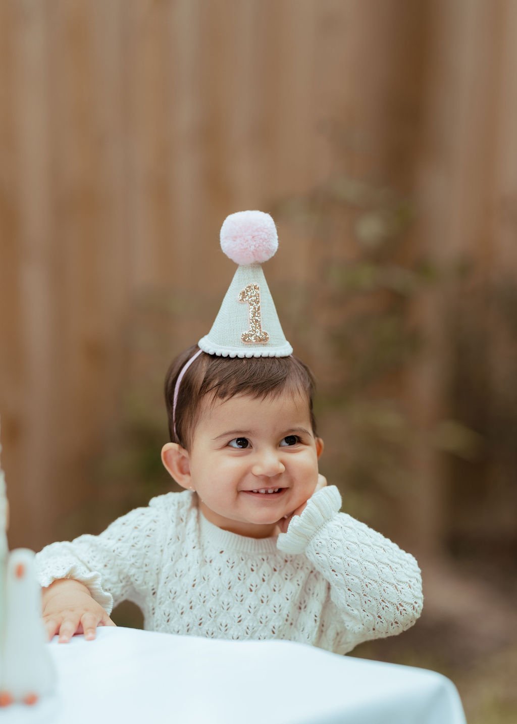 A young child with short dark hair, smiling and wearing a white knit sweater, sitting at a table with a birthday party hat on her head. The hat has a number '1' and a pink pompom on top. The background is blurred.
