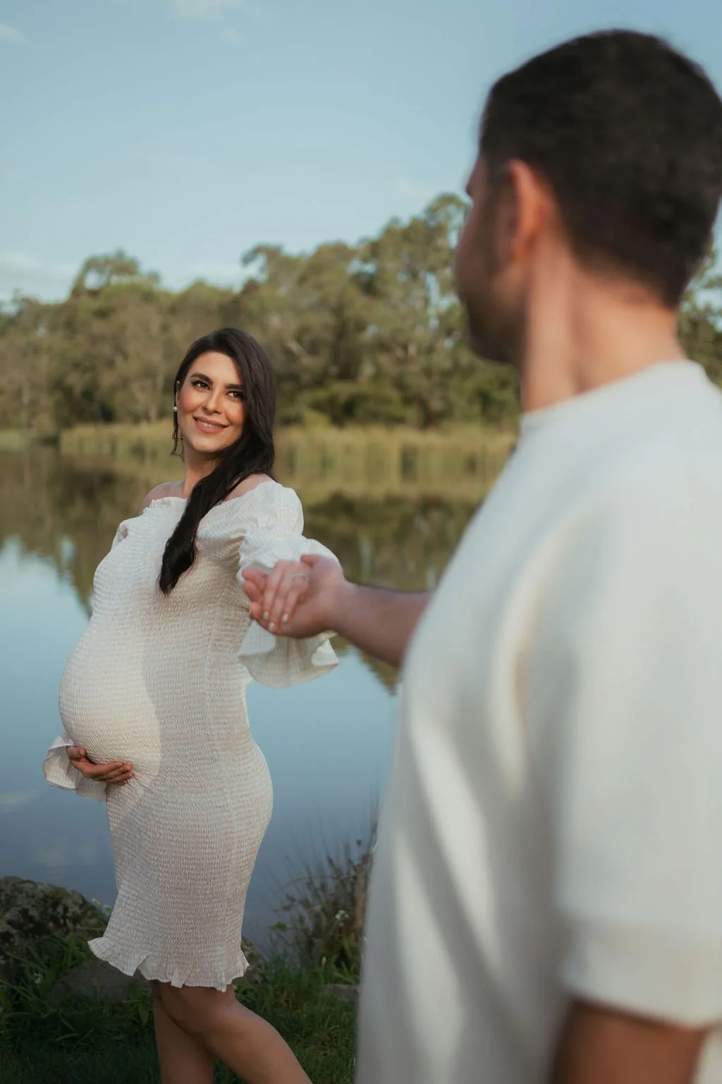 A pregnant woman in a white dress smiling and holding her belly, standing by a lake, while holding hands with a man whose face is not visible, in a scenic outdoor setting with trees and water.