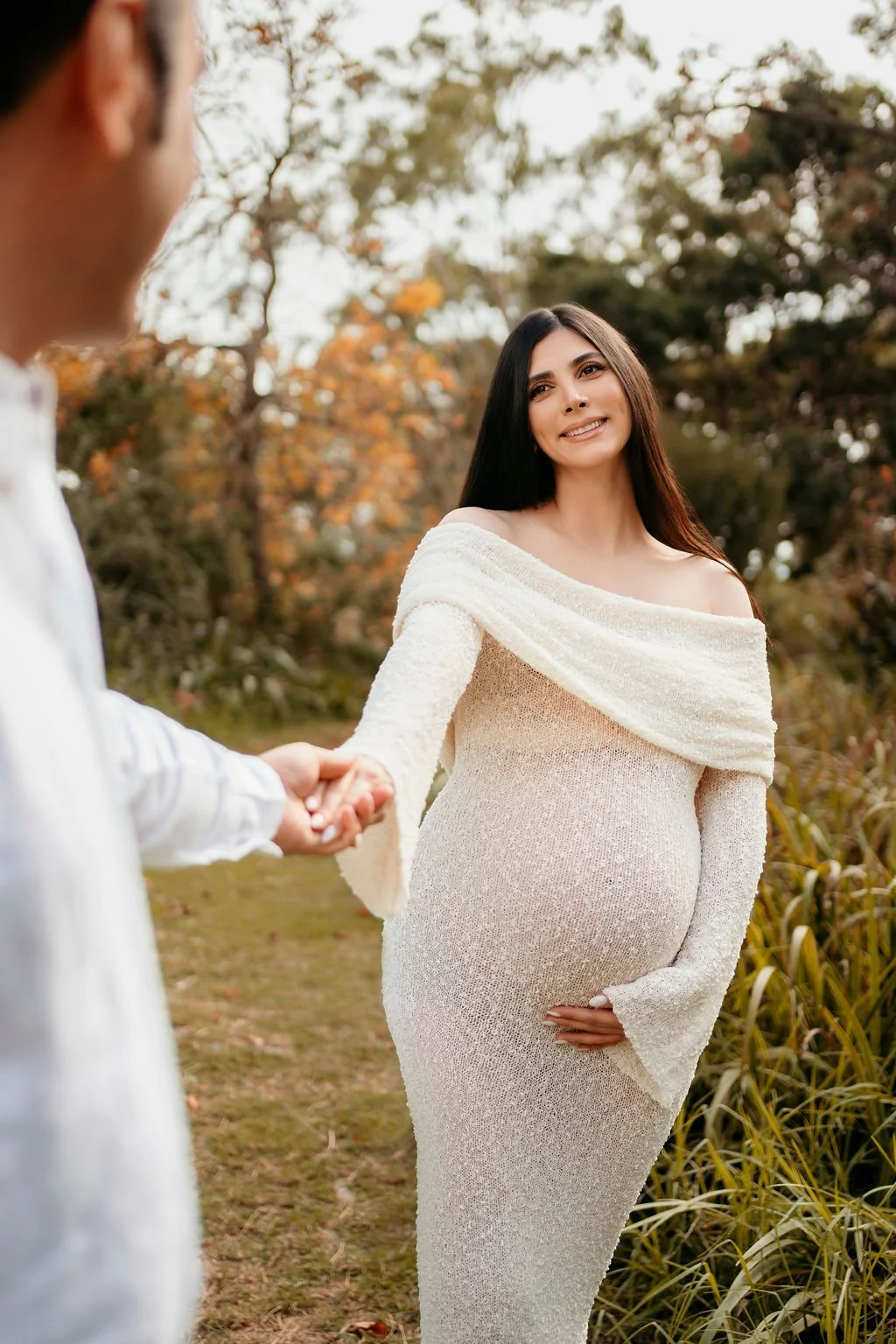 A pregnant woman in a light-colored dress stands outdoors in a natural setting, holding her belly with one hand and smiling at a man who is holding her hand.