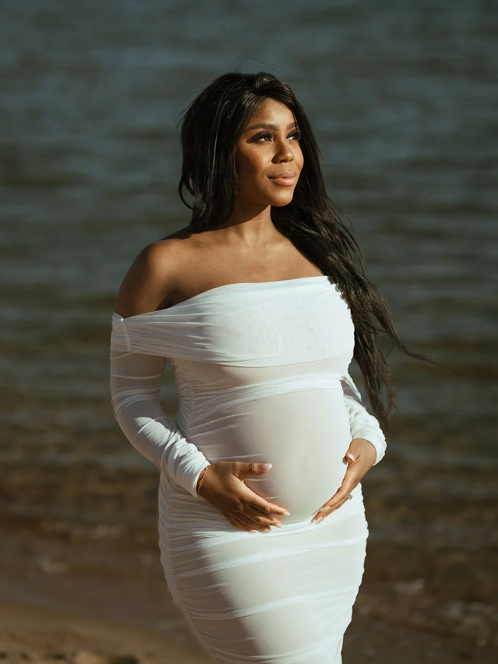 Pregnant woman in a white off-the-shoulder dress standing on a beach, cradling her belly and looking thoughtfully into the distance near the water.