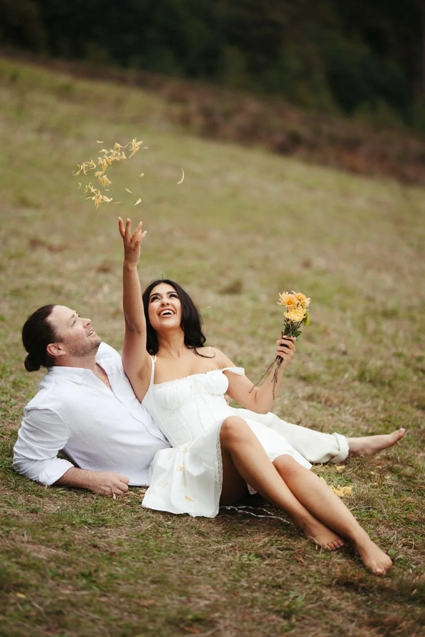 A couple sitting on the grass outdoors, with the woman throwing flower petals into the air and holding a bouquet, both smiling and enjoying each other's company.