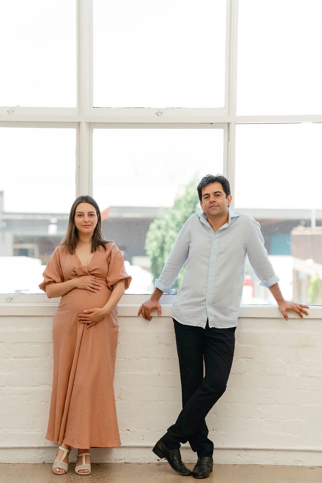 A pregnant woman in a brown dress with her hand on her belly, standing next to a man in a white shirt with rolled-up sleeves and black pants, leaning against a windowsill in front of a large window with a view of buildings and trees.
