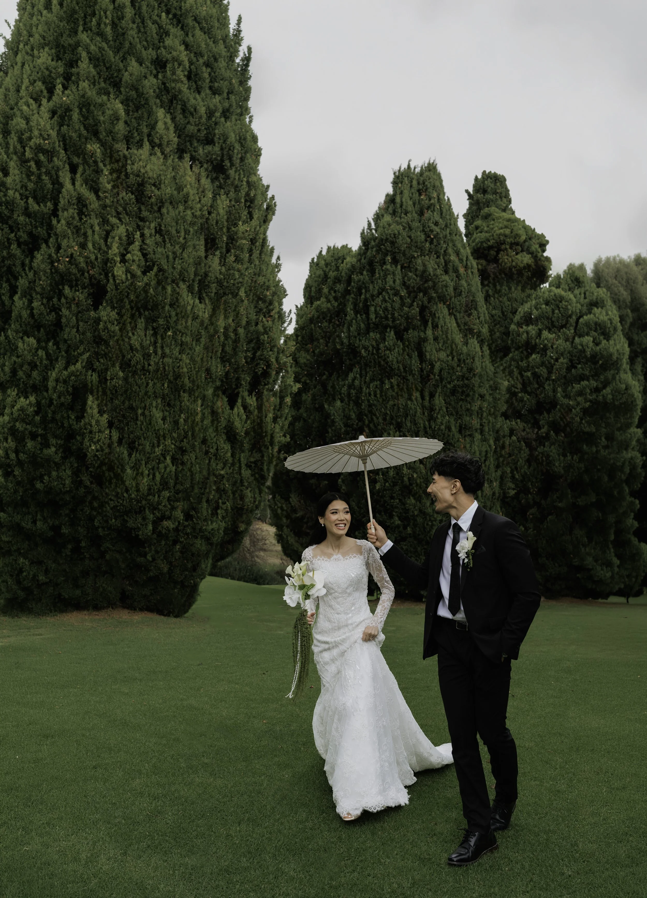 Woman smiling and wearing a white dress, walking outdoors through greenery, with another person partially visible in the background.