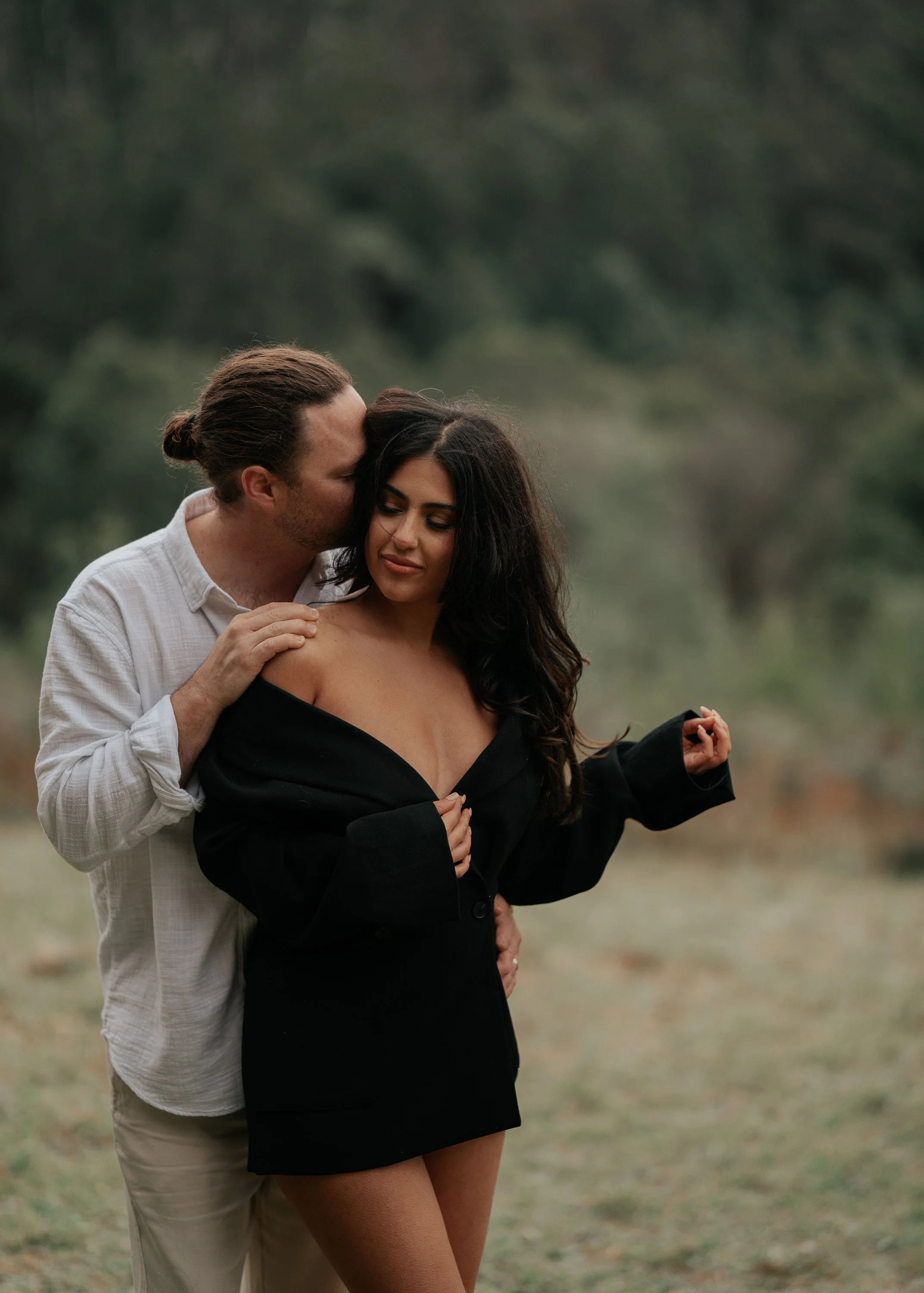 A man and a woman standing outdoors in a grassy area, with the man kissing the woman's forehead while she closes her eyes and has a soft smile, wearing a black off-shoulder top and the man wearing a white shirt.
