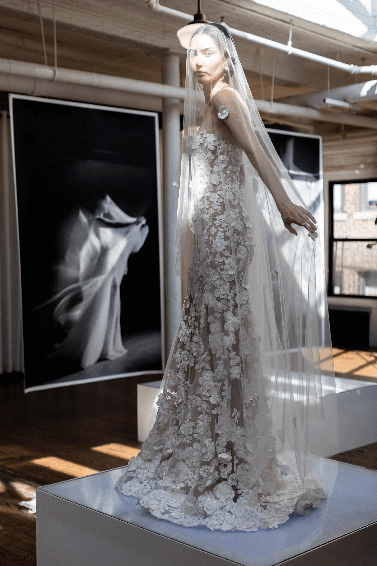 A woman wearing a sheer lace wedding dress with floral appliqués, standing on a white pedestal in a gallery or studio.
