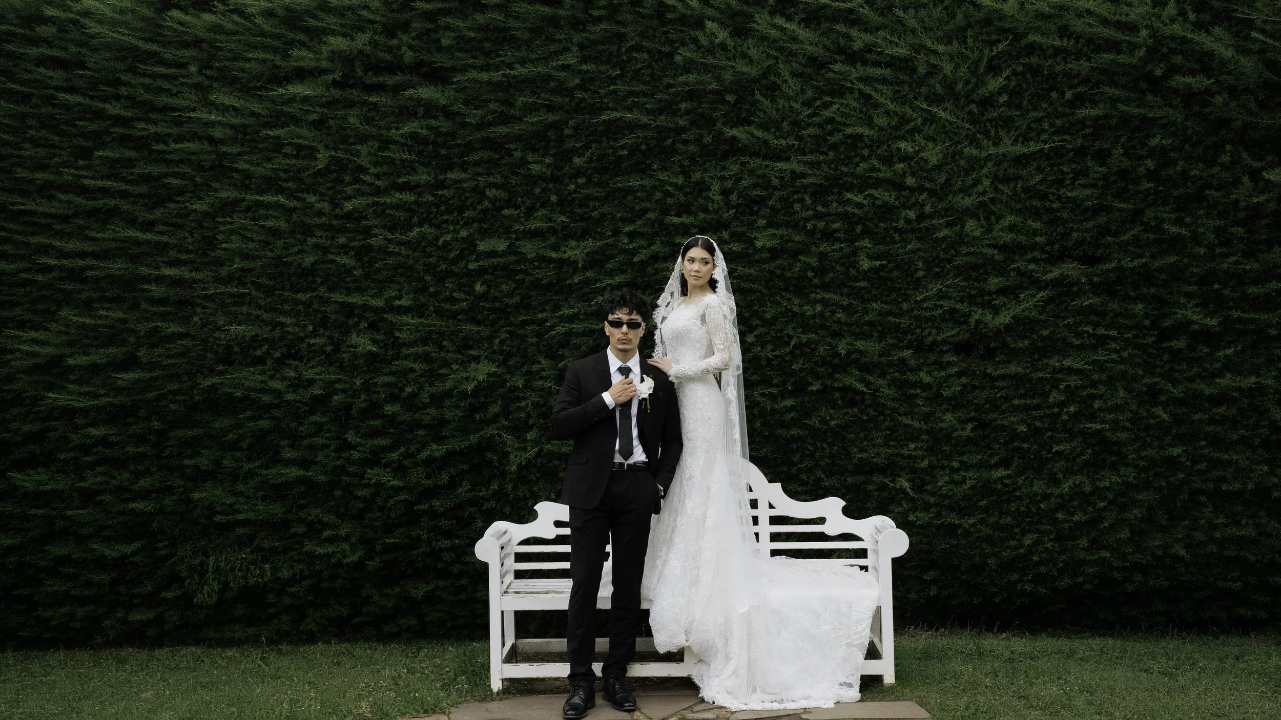 A bride and groom in wedding attire pose in front of a large green hedge, with the bride standing on a white bench and the groom kneeling beside her, holding her hand, during a wedding photoshoot.