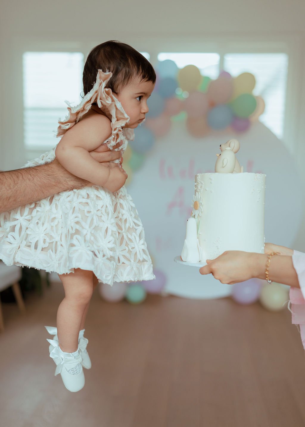 A young girl in a white, floral dress is being held up by an adult. She is bending forward and looking at a birthday cake with animal decorations. Someone is holding the cake on a tray, preparing to give it to her during a celebration with colorful b