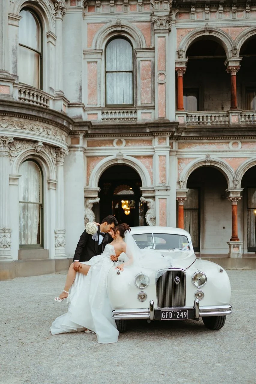 a bride and groom kissing in front of a vintage white car outside a grand historic building