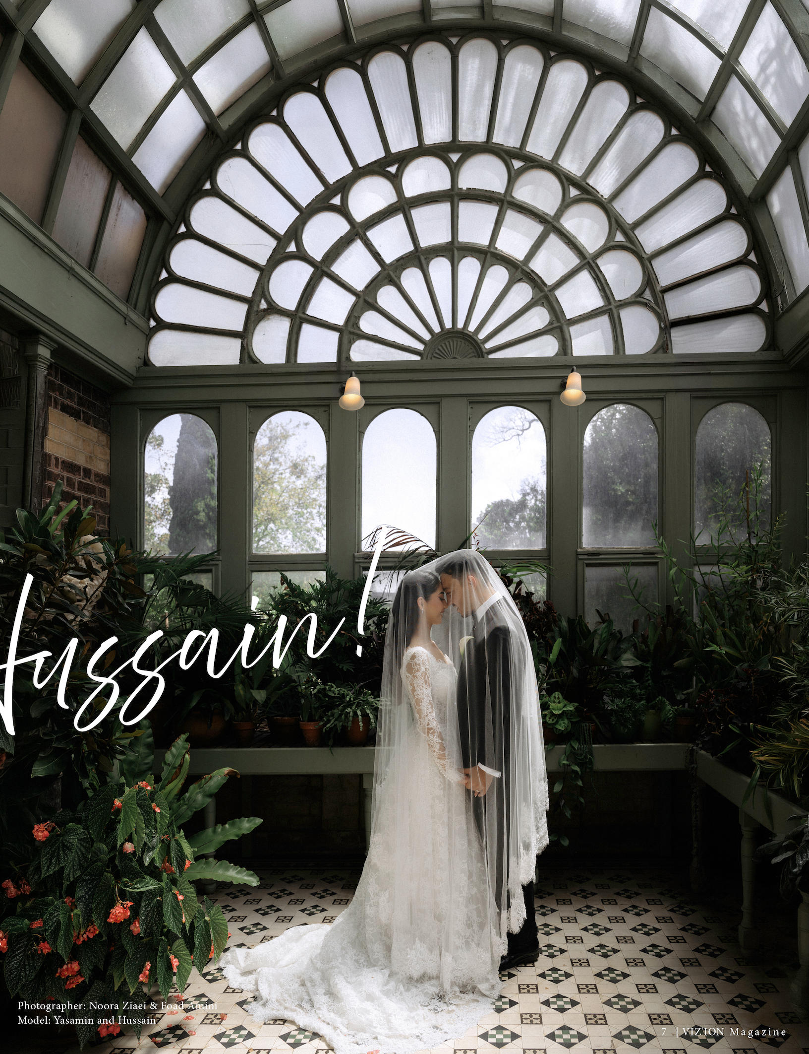 A bride and groom standing close together, touching foreheads, in a glass conservatory with a large decorative arch window above them and lush plants surrounding them.