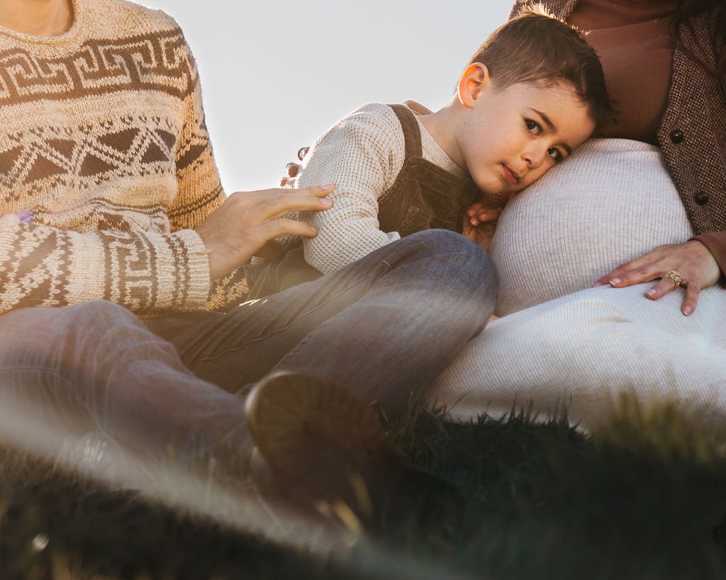 A young boy leaning his head on a pregnant woman's belly while sitting outdoors on the grass, with another person touching his arm, during sunset.