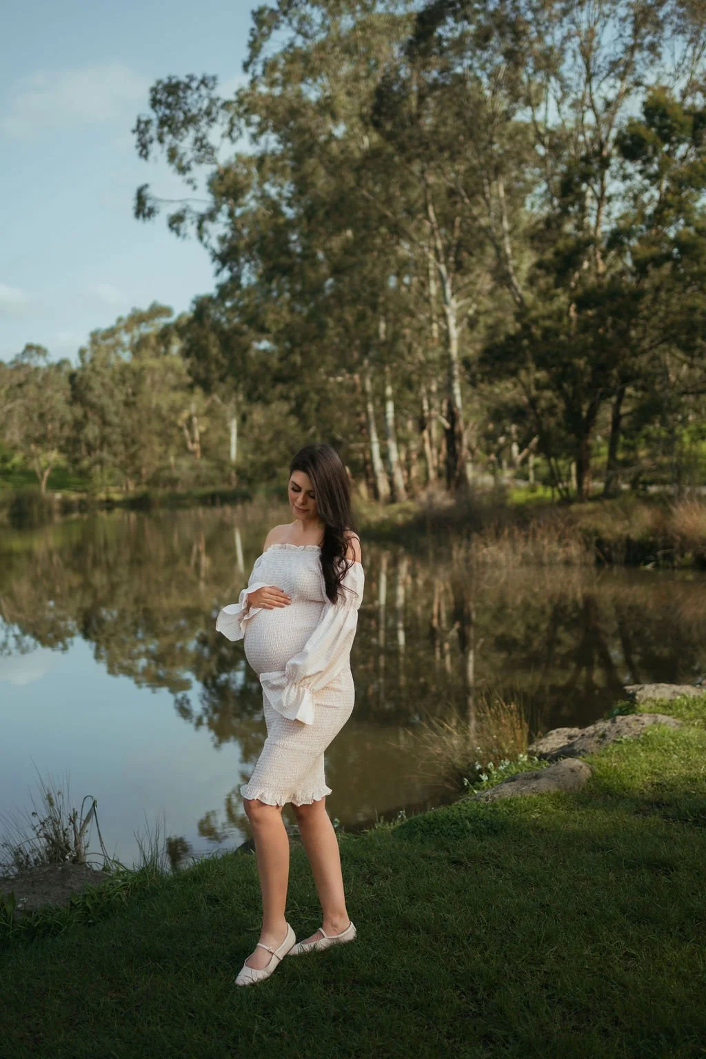 Pregnant woman in white dress standing near a lake with trees in the background.