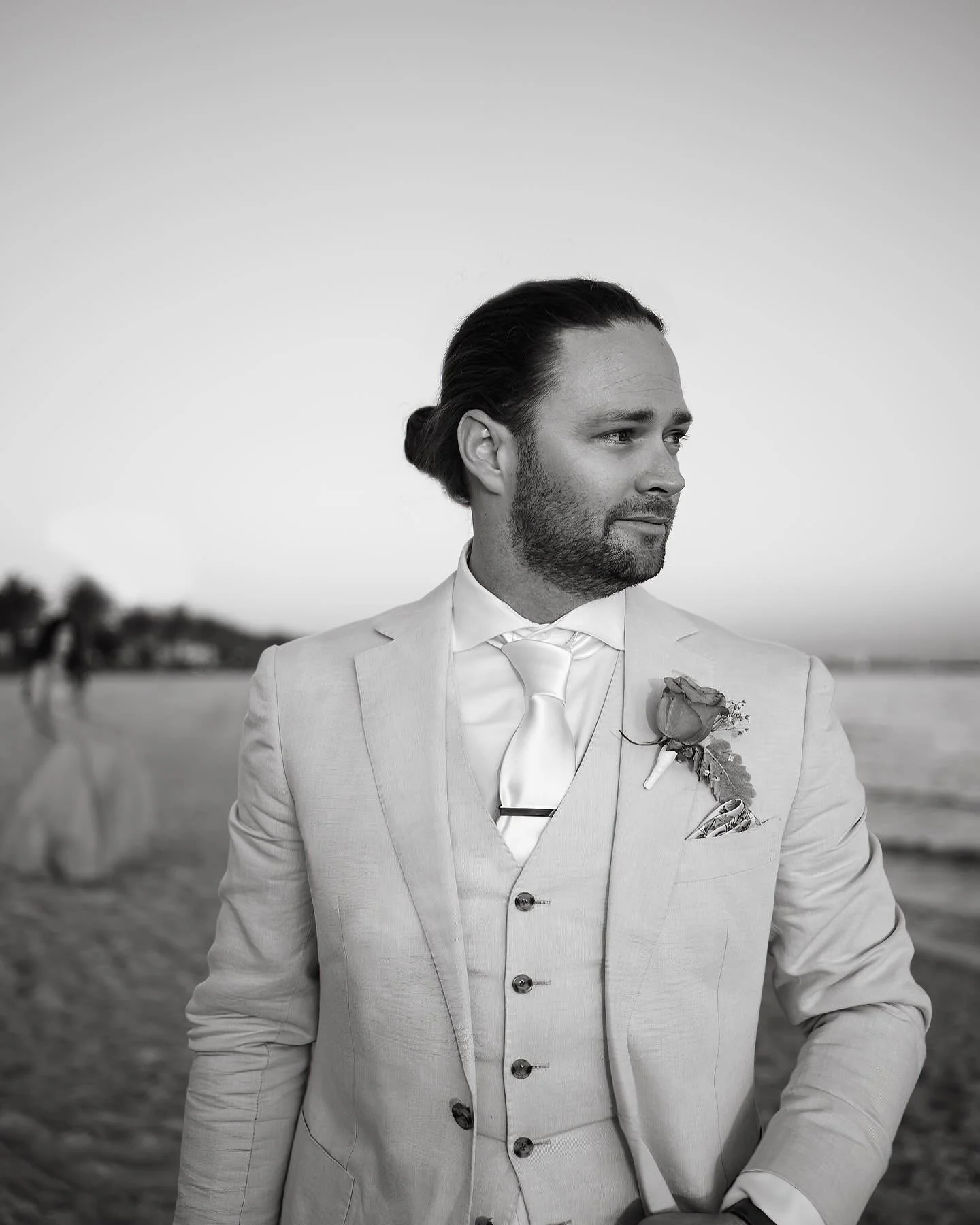 A man in a light-colored suit, vest, and tie looking to the right, standing on a beach with blurred figures in the background.