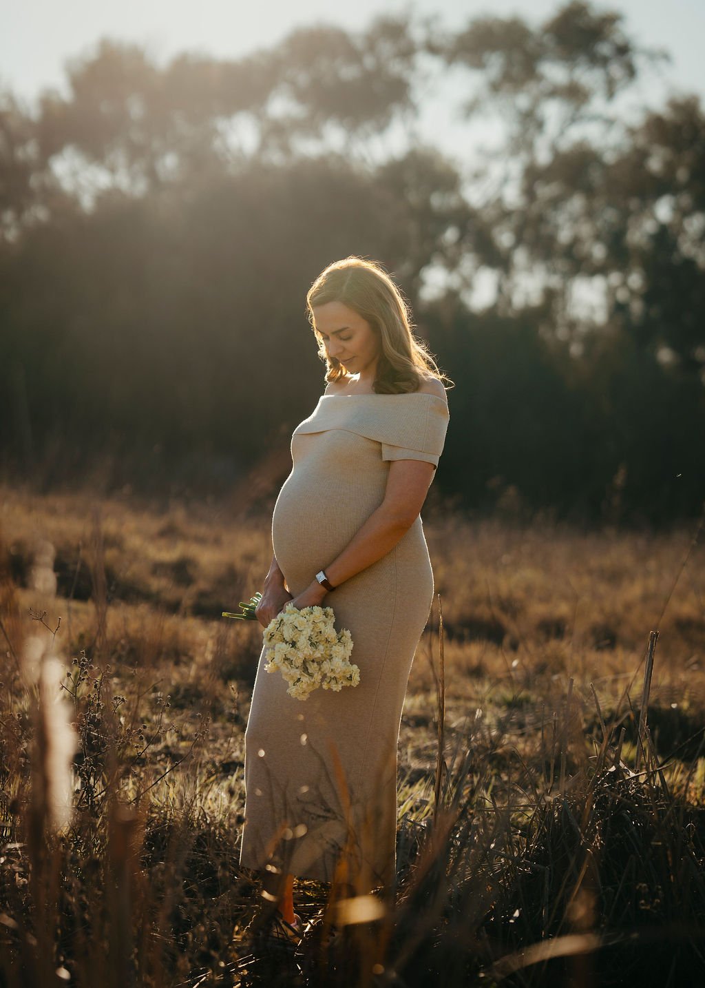 A pregnant woman in a beige off-shoulder dress standing in a field during sunset, holding a bouquet of white flowers, with a serene expression on her face.