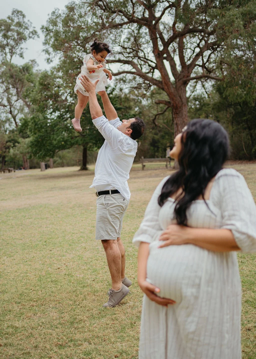 A man is lifting a young girl into the air in a park, while a pregnant woman watches and smiles nearby. Trees are in the background.
