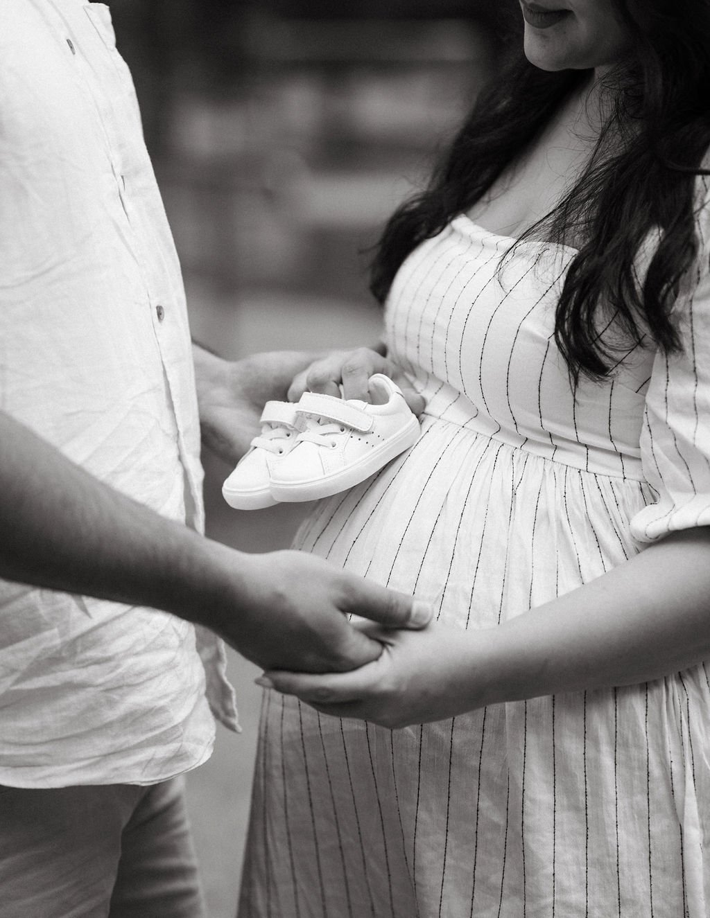 A pregnant woman holds a pair of baby shoes with a man standing in front of her, holding her hands.